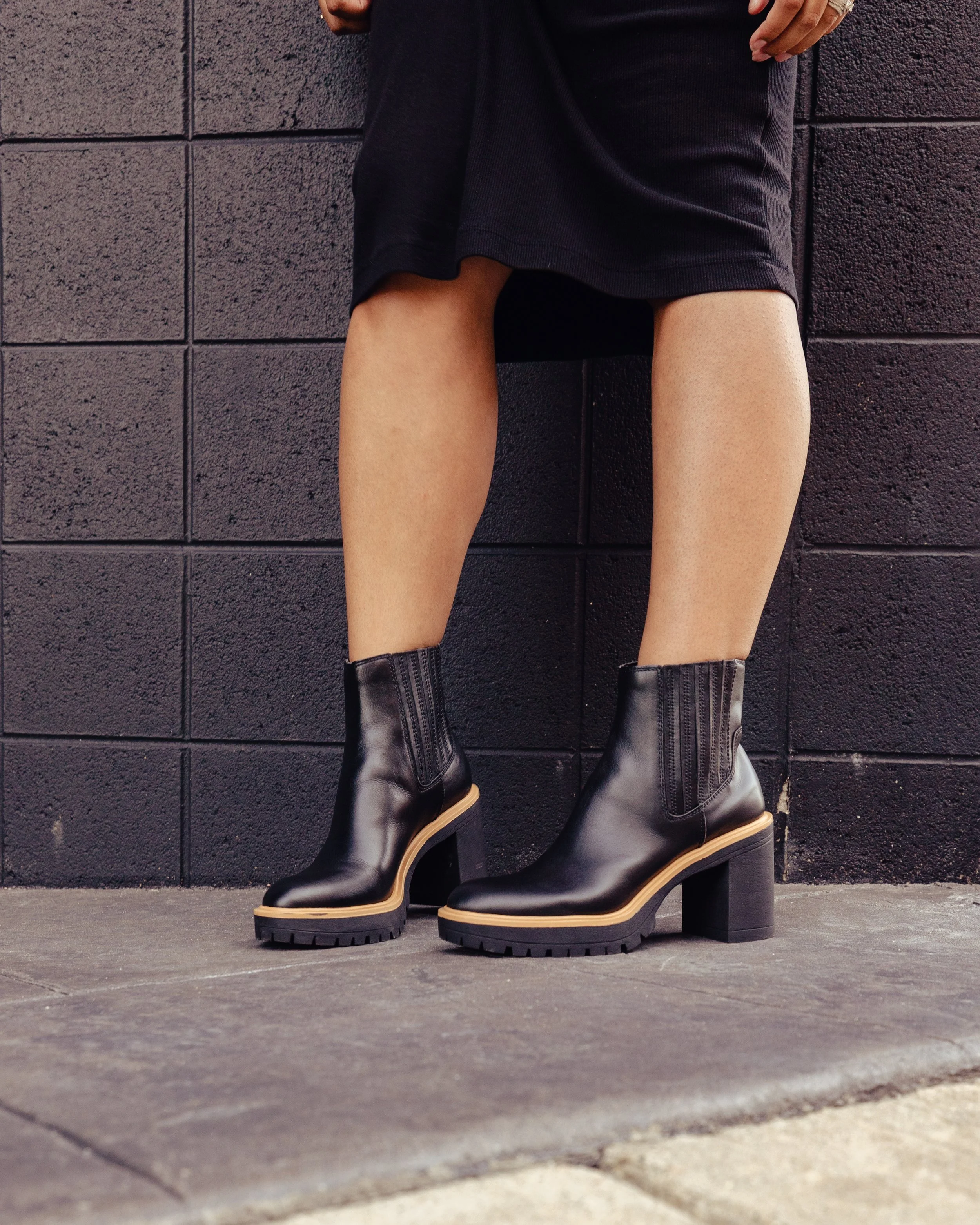 Close-up of a person wearing black leather ankle boots with chunky heels, standing against a black brick wall, with a black skirt or dress.