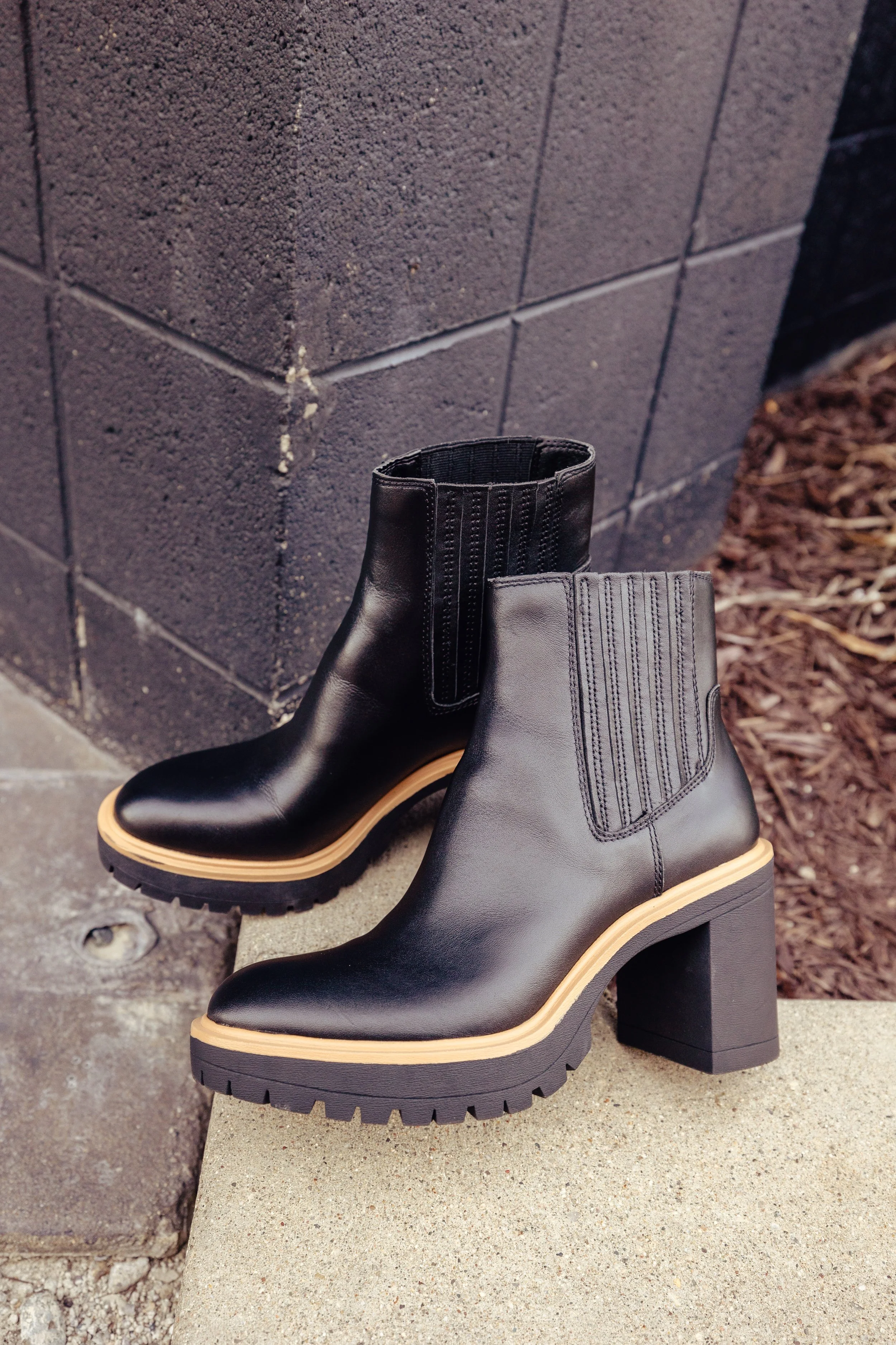 Black leather high-heeled ankle boots with tan soles and elastic side panels, placed on a concrete surface near a dark brick wall.