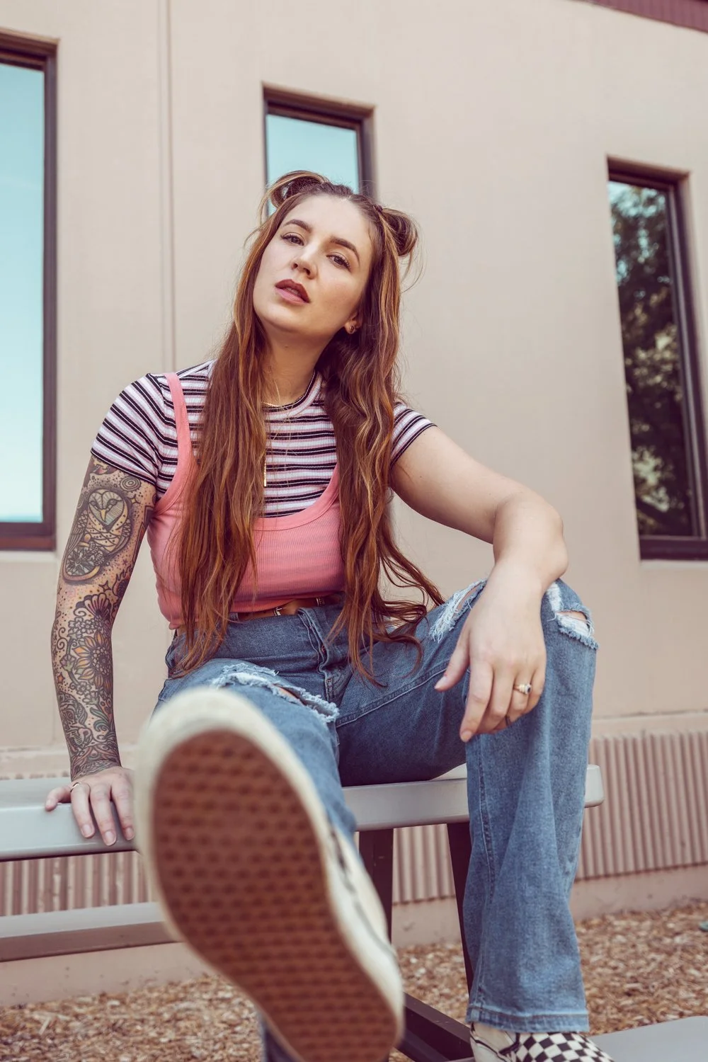 A young woman with long red hair, tattoos on her arm, wearing a striped shirt and pink tank top, sitting on a bench outside a modern building, looking at the camera with a casual pose and her foot close to the lens.