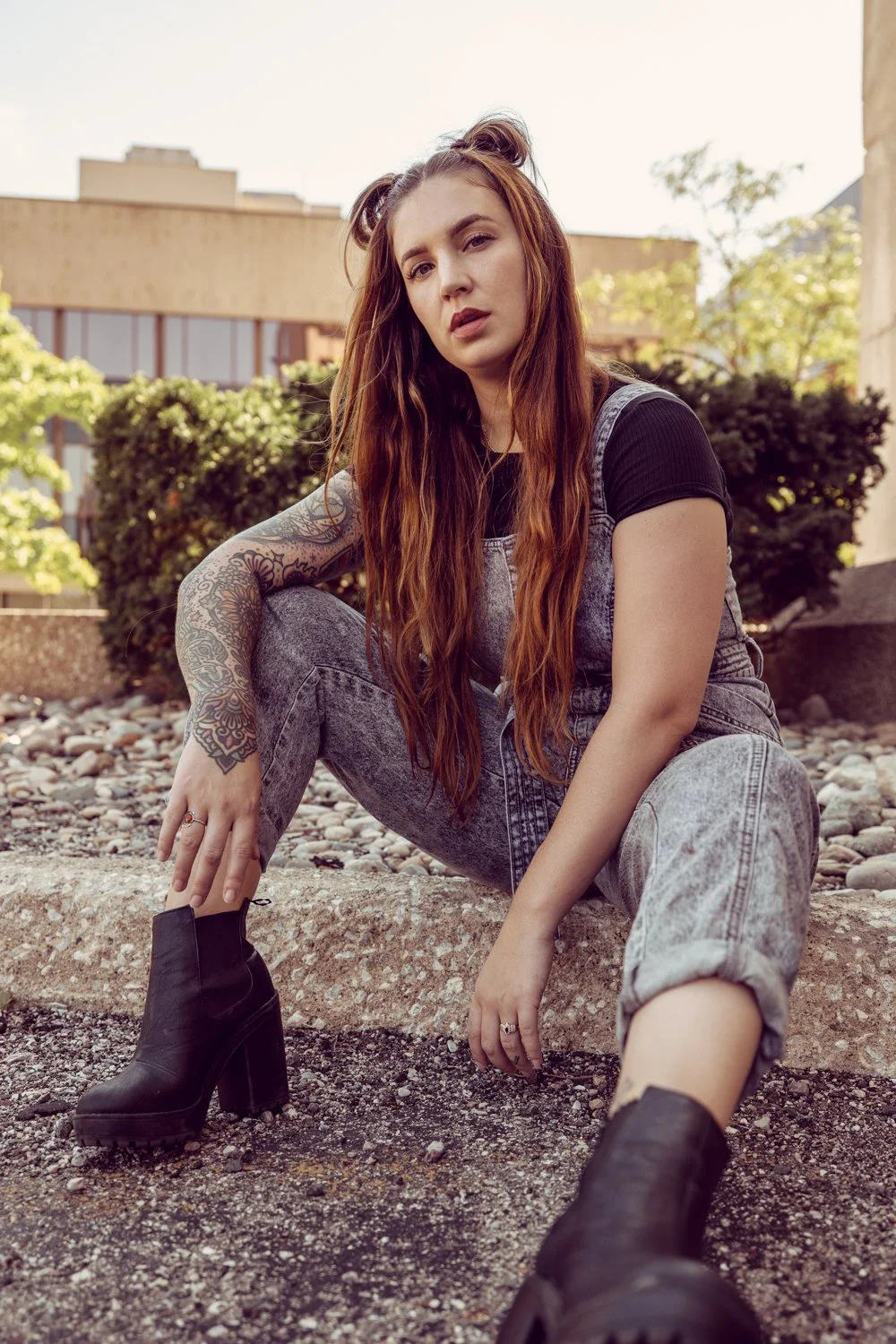 A young woman with long red hair, sitting on a stone ledge outdoors, wearing a black t-shirt, faded jeans, and black high-heeled boots, with a tattooed arm and a serious expression.