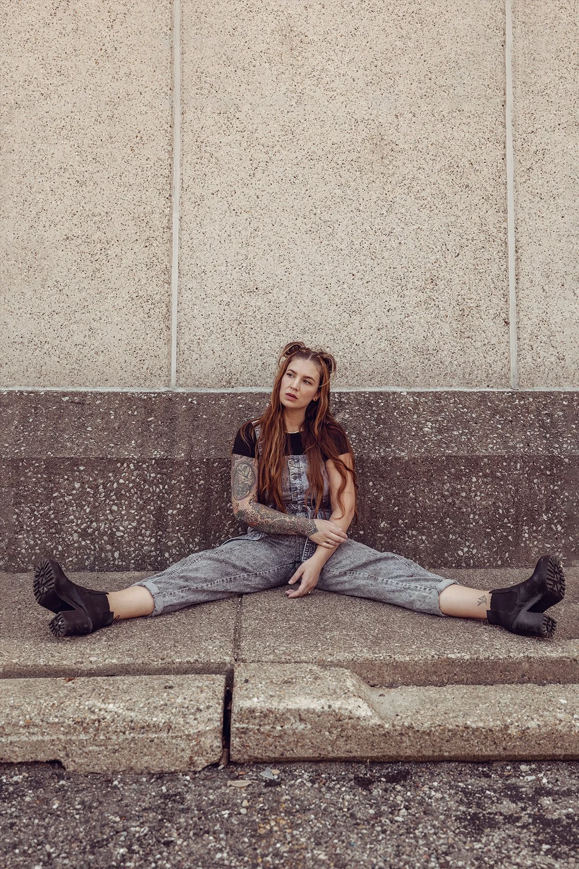 A young woman with tattoos sitting on a sidewalk, leaning against a wall, with legs in a split position.
