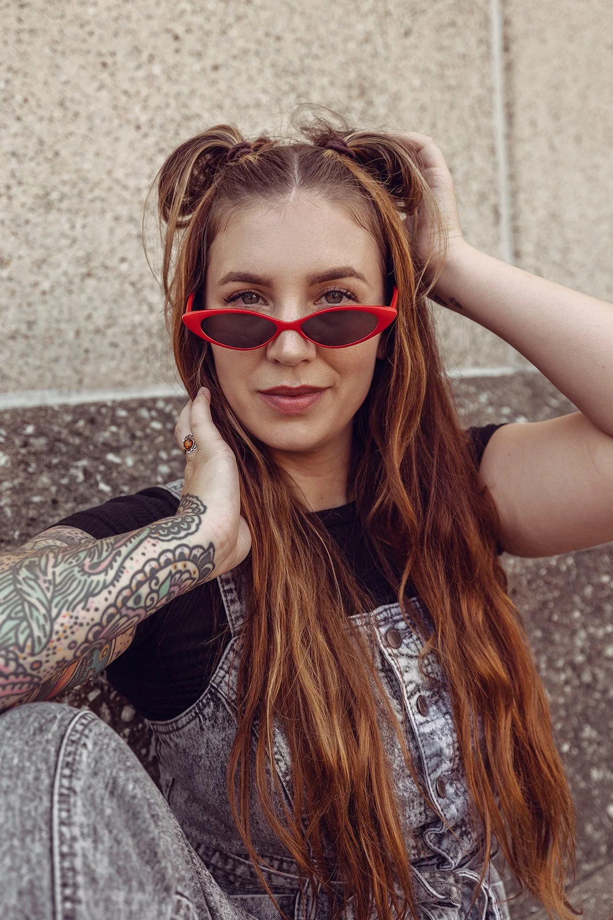 A young woman with long red hair, wearing red sunglasses, a black top, and gray denim overalls, sitting outside against a textured wall.