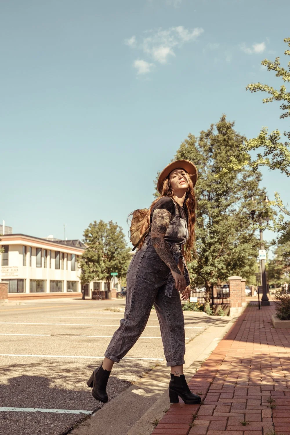 A woman with long red hair wearing a hat, black ankle boots, and overalls stands on a sidewalk in an urban area with trees and buildings in the background, looking up.