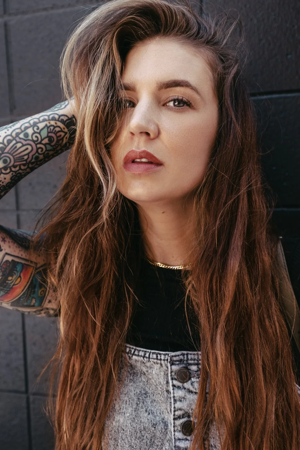 A young woman with long wavy brown hair, tattoos on her arms, wearing a black top, a chain necklace, and light-colored jeans, standing against a dark brick wall.
