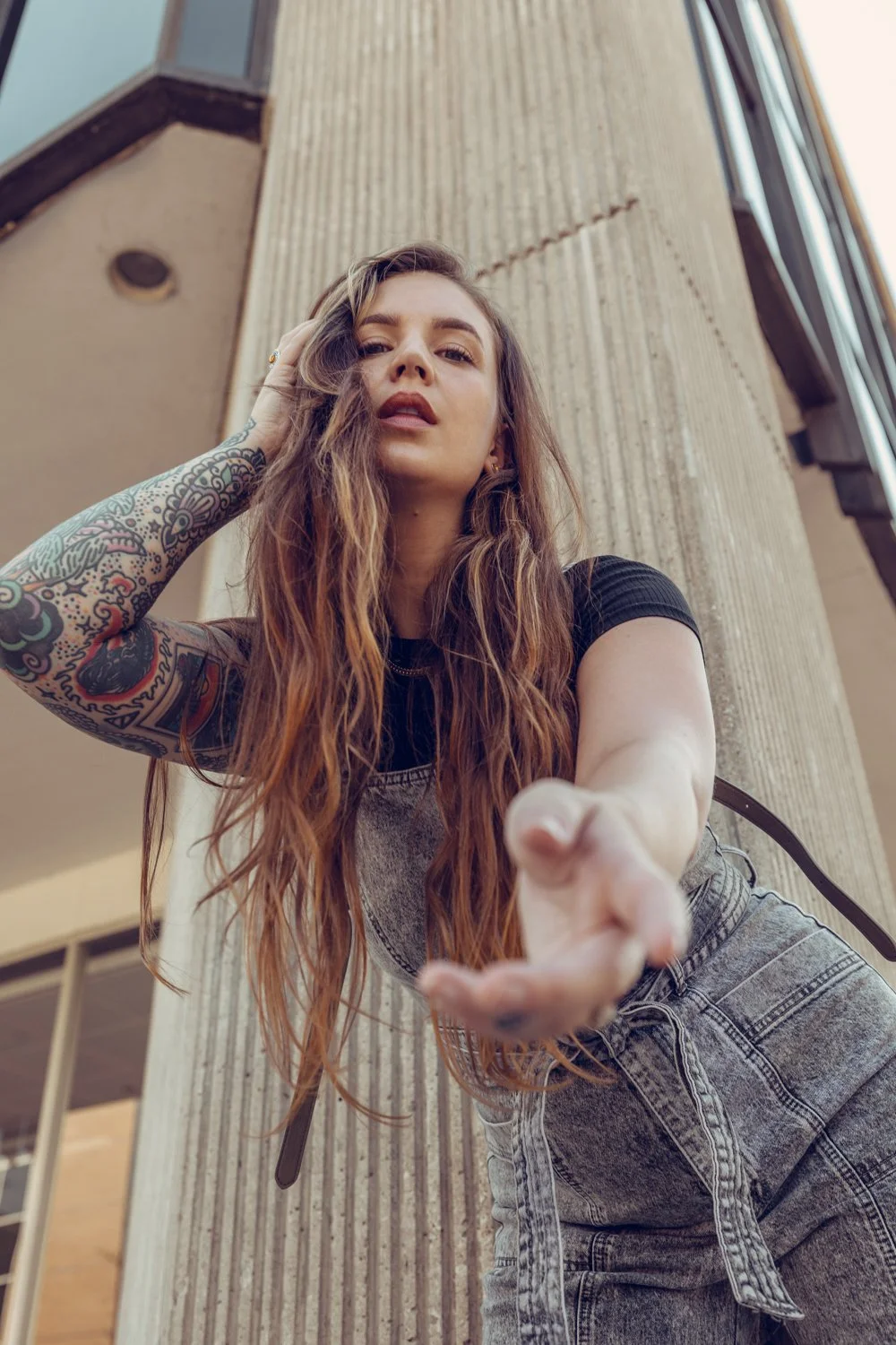 A woman with long wavy hair and tattoos on her right arm with an upward-looking camera angle showing a building with vertical concrete panels.