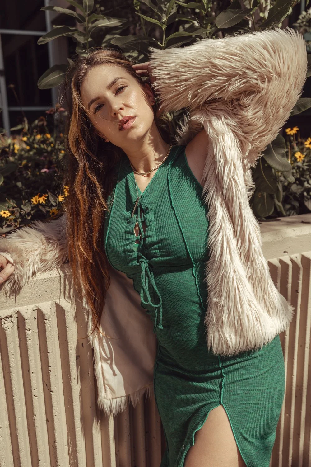 A woman with long, wavy hair and light makeup, posing outdoors near a brick wall and plants. She wears a green dress with a slit and a beige coat with faux fur trim, holding her hair back with one arm.