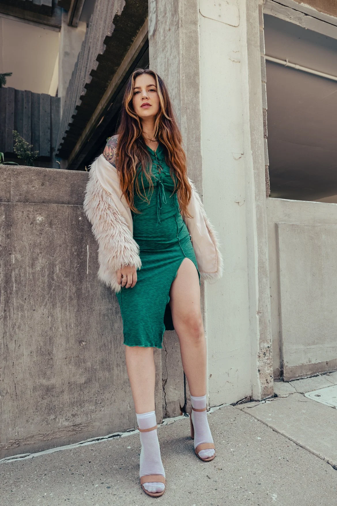 Young woman with long wavy hair wearing a green dress with a high slit, a cream fur jacket, and white high-heeled shoes standing against a concrete wall in an urban setting.