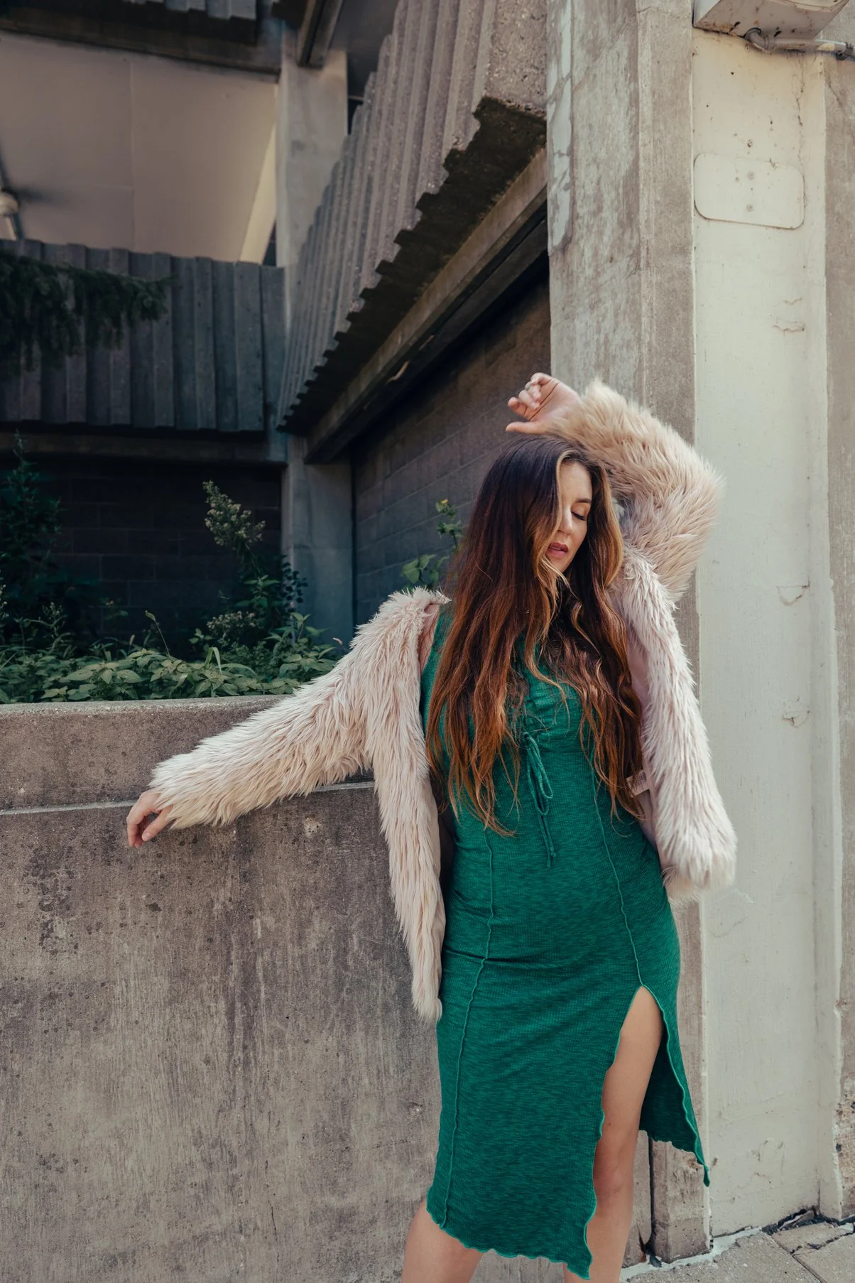 A woman with long wavy hair wearing a green dress with a high slit and a fluffy beige jacket, standing outdoors against a concrete wall with her eyes closed and arm raised.