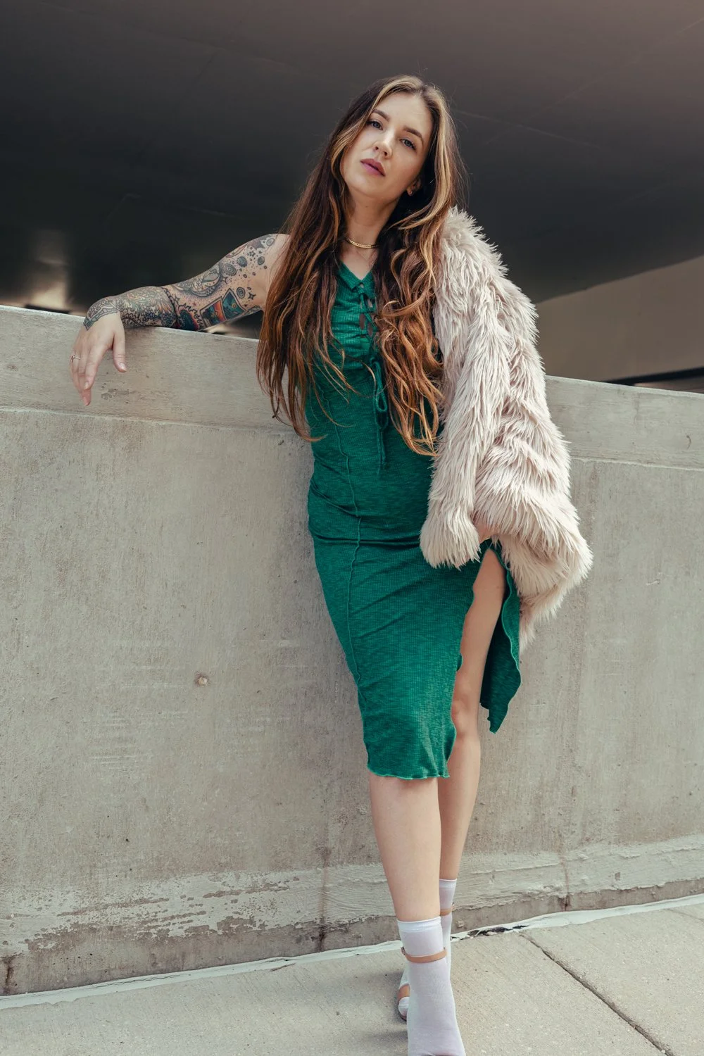 A young woman with long brown hair and tattoos on her arm leaning on a concrete wall. She is wearing a green dress, white socks, and a beige faux fur jacket in an urban setting.