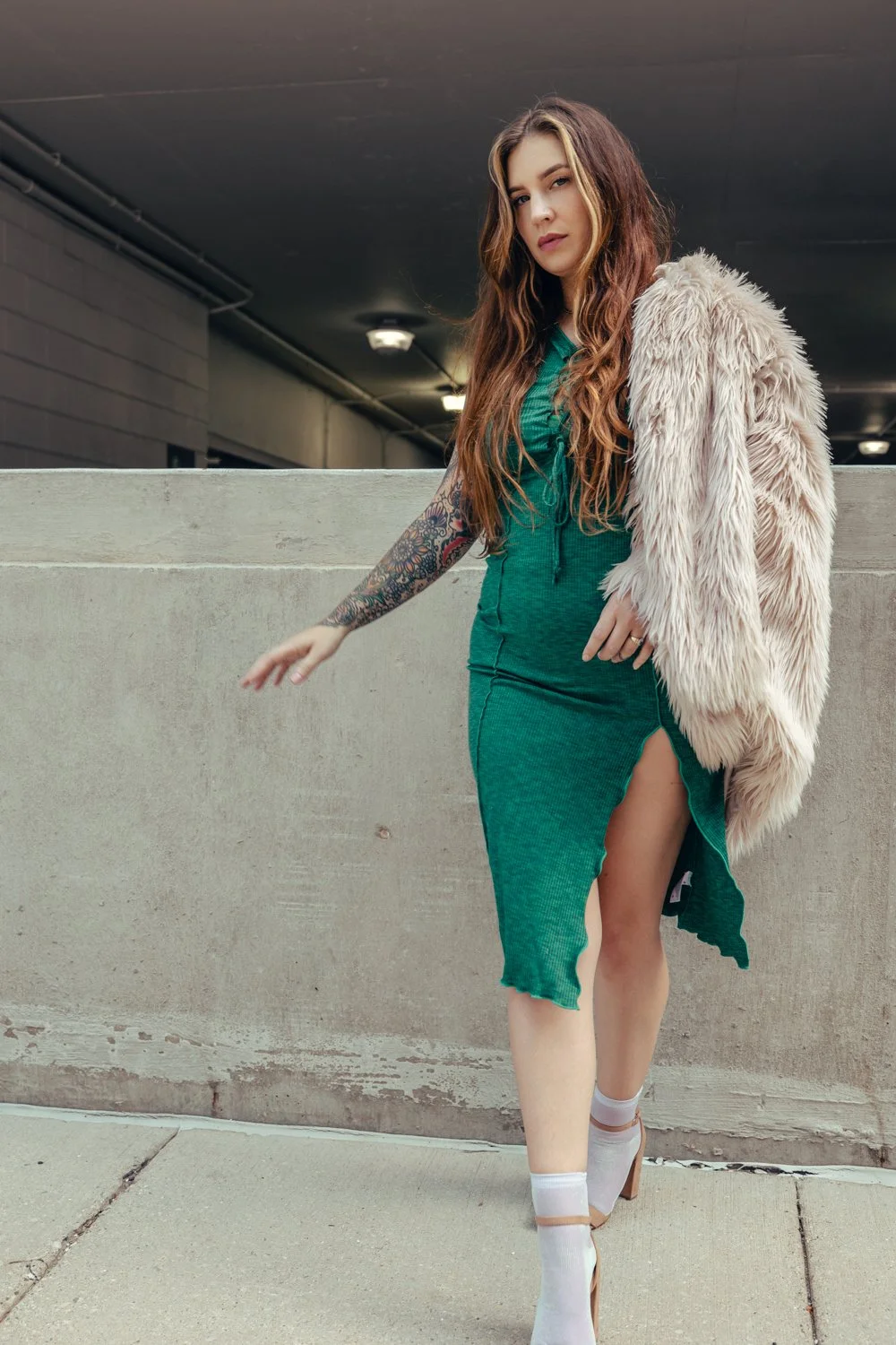 A woman with long wavy hair, wearing a teal dress with a slit and a fluffy cream-colored jacket, stands against a concrete wall in an urban setting at night.