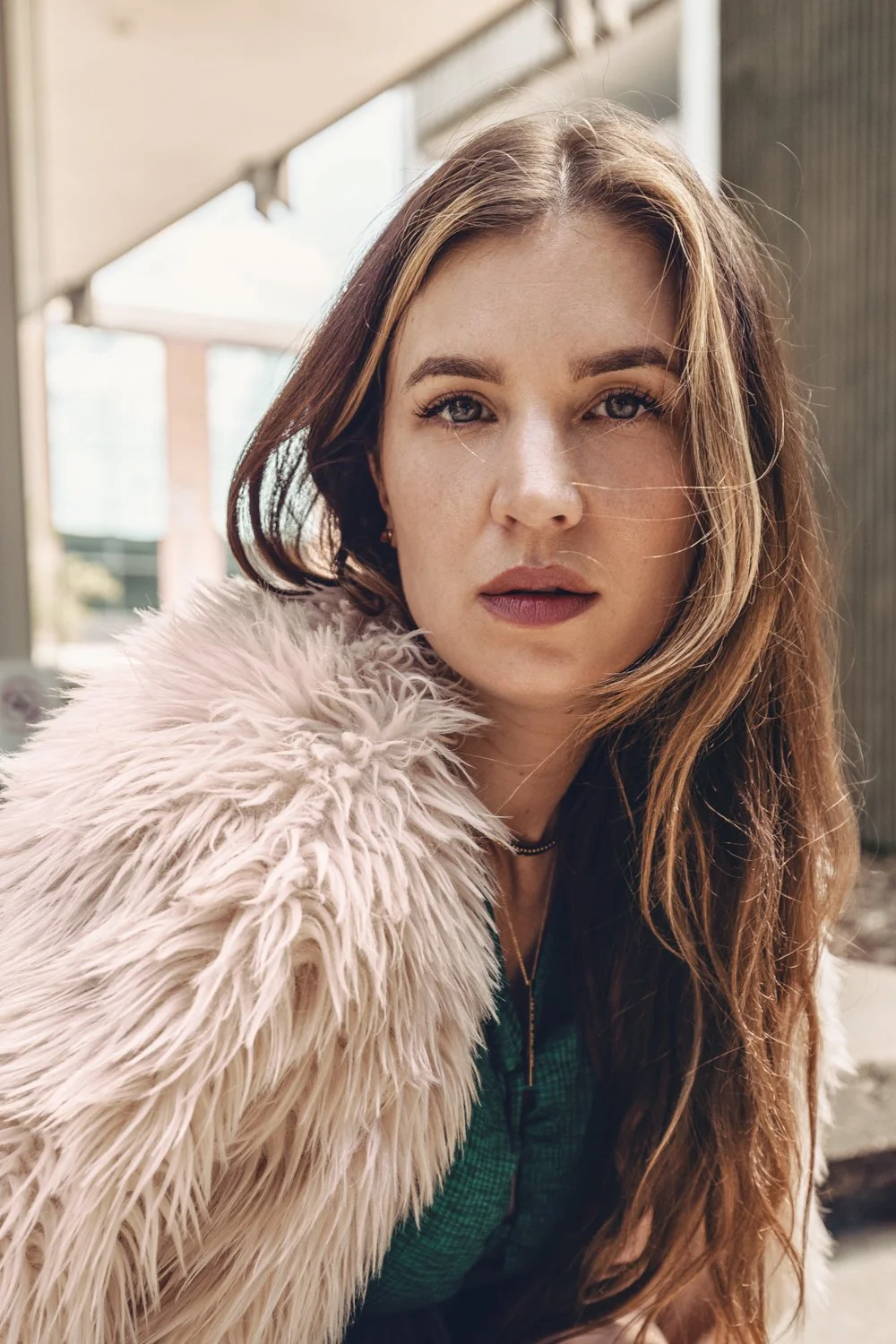 A woman with long brown hair and blue eyes wearing a pink fluffy coat outdoors.