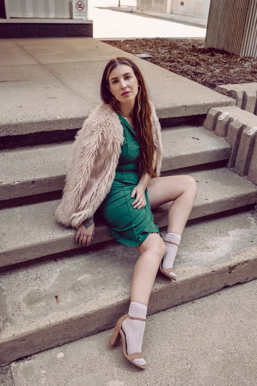 A woman with long brown hair in a green dress and high heels, sitting on steps outdoors, wearing a pink faux fur coat over her shoulders.