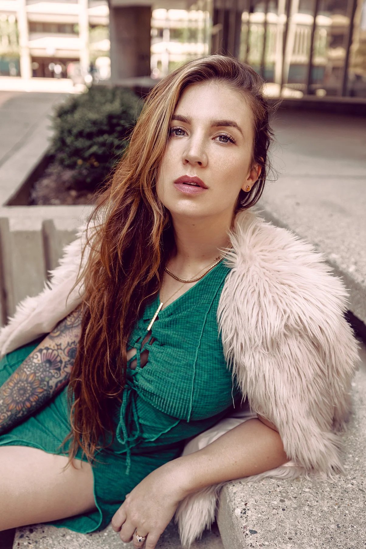 A woman with long red hair, wearing a green dress and a fluffy pink coat, sitting outdoors on a concrete ledge with buildings and greenery in the background.
