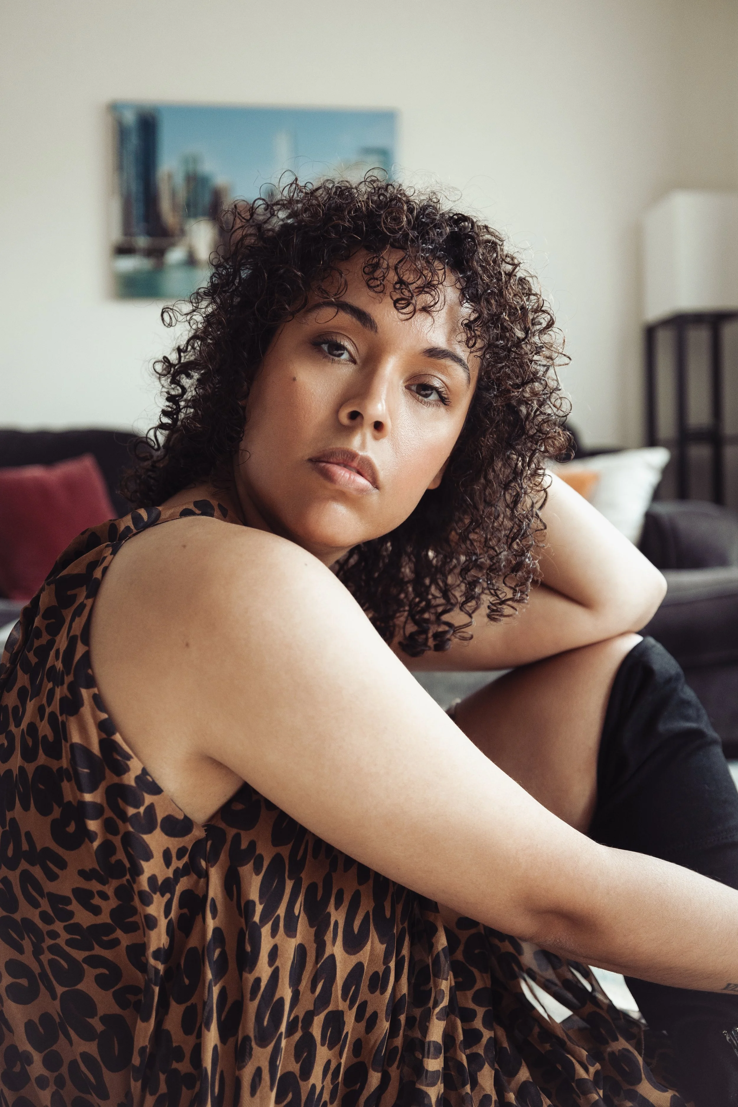 A woman with curly hair sitting on a couch, looking directly at the camera. She is wearing a leopard print sleeveless top.
