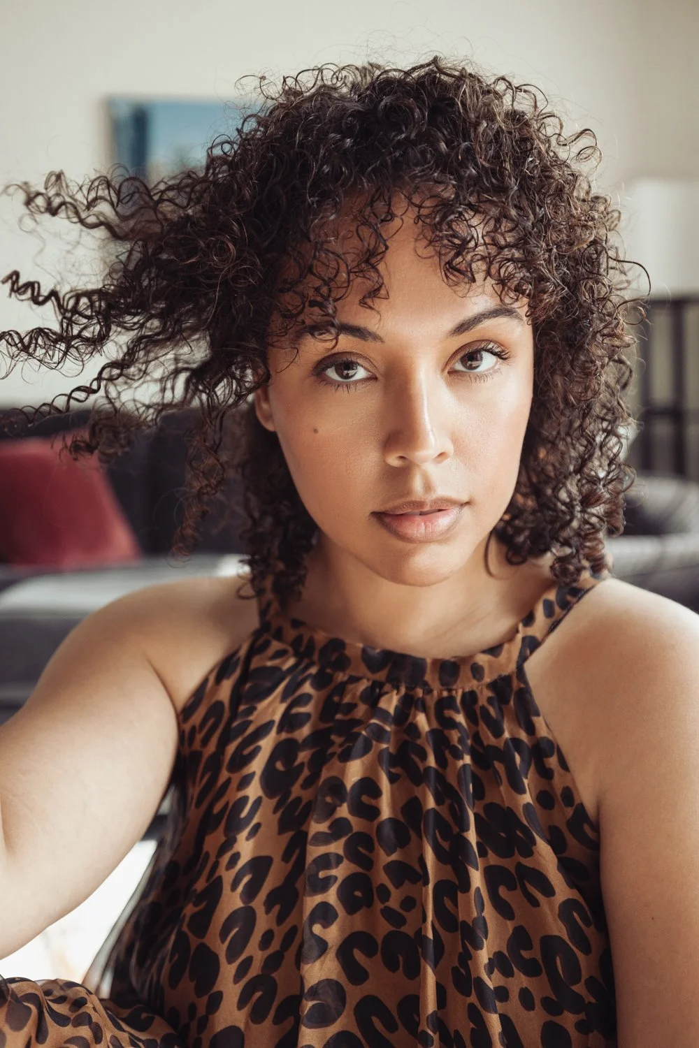 A woman with curly hair wearing a leopard print top, looking at the camera in a room with a couch and framed artwork in the background.