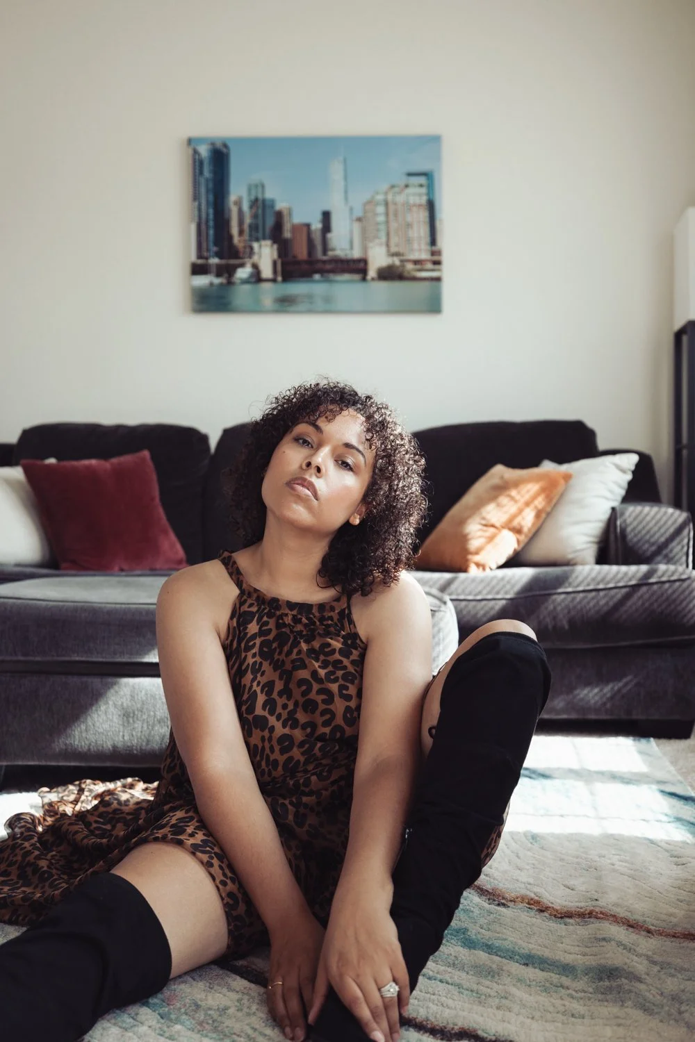 A woman with curly hair sitting on the floor in front of a gray sofa with multiple pillows, wearing a leopard print dress and black boots, in a living room with a large cityscape picture hanging on the wall.