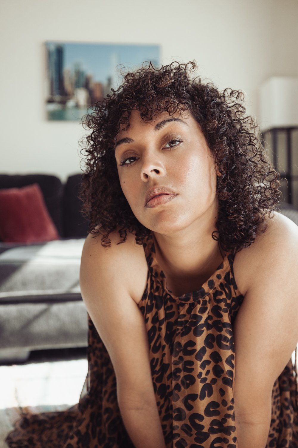 A woman with curly dark hair and a leopard print top, looking directly at the camera, in a living room setting.