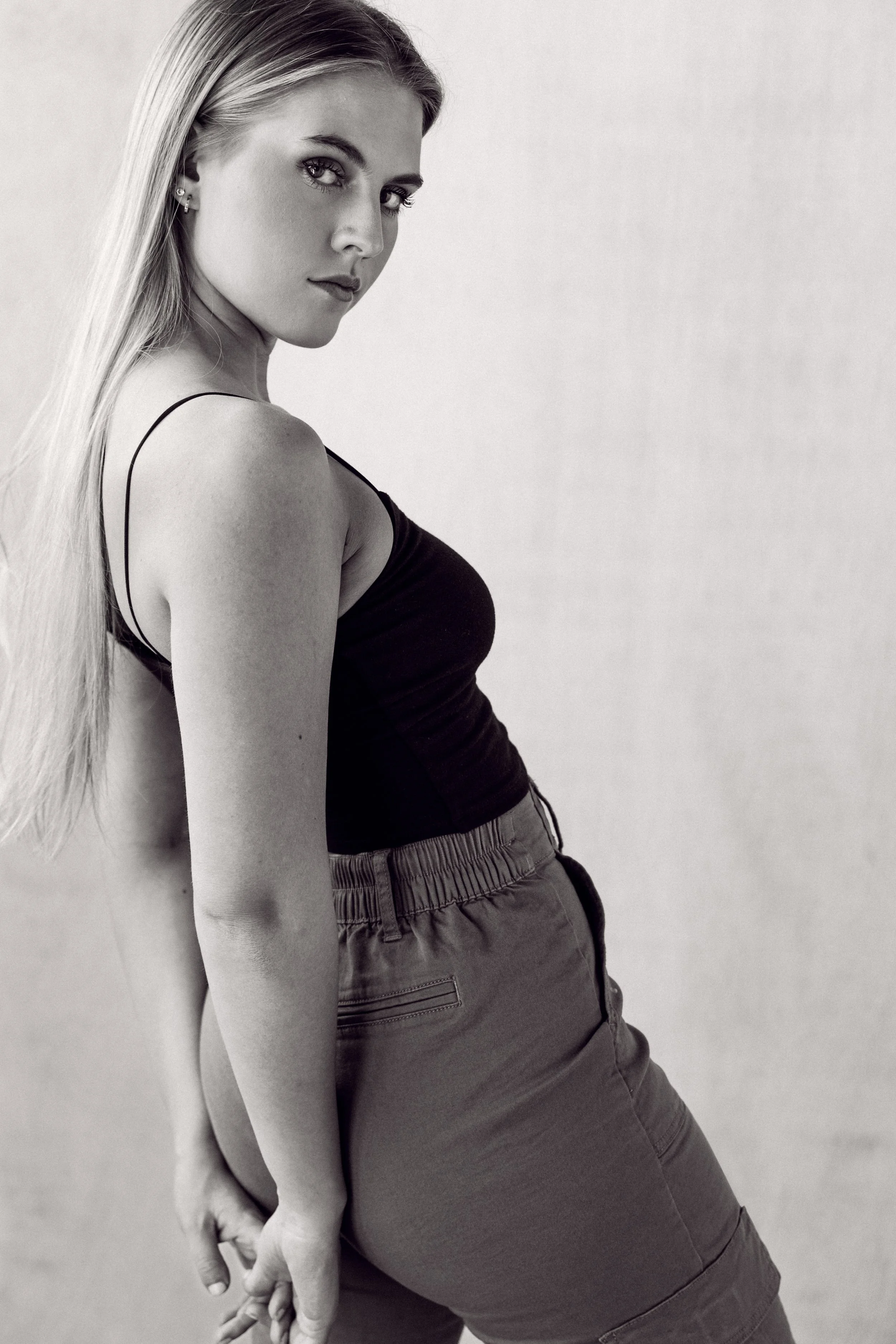 A young woman with long, straight blonde hair and striking eyes, wearing a black tank top and casual shorts, standing against a plain background.