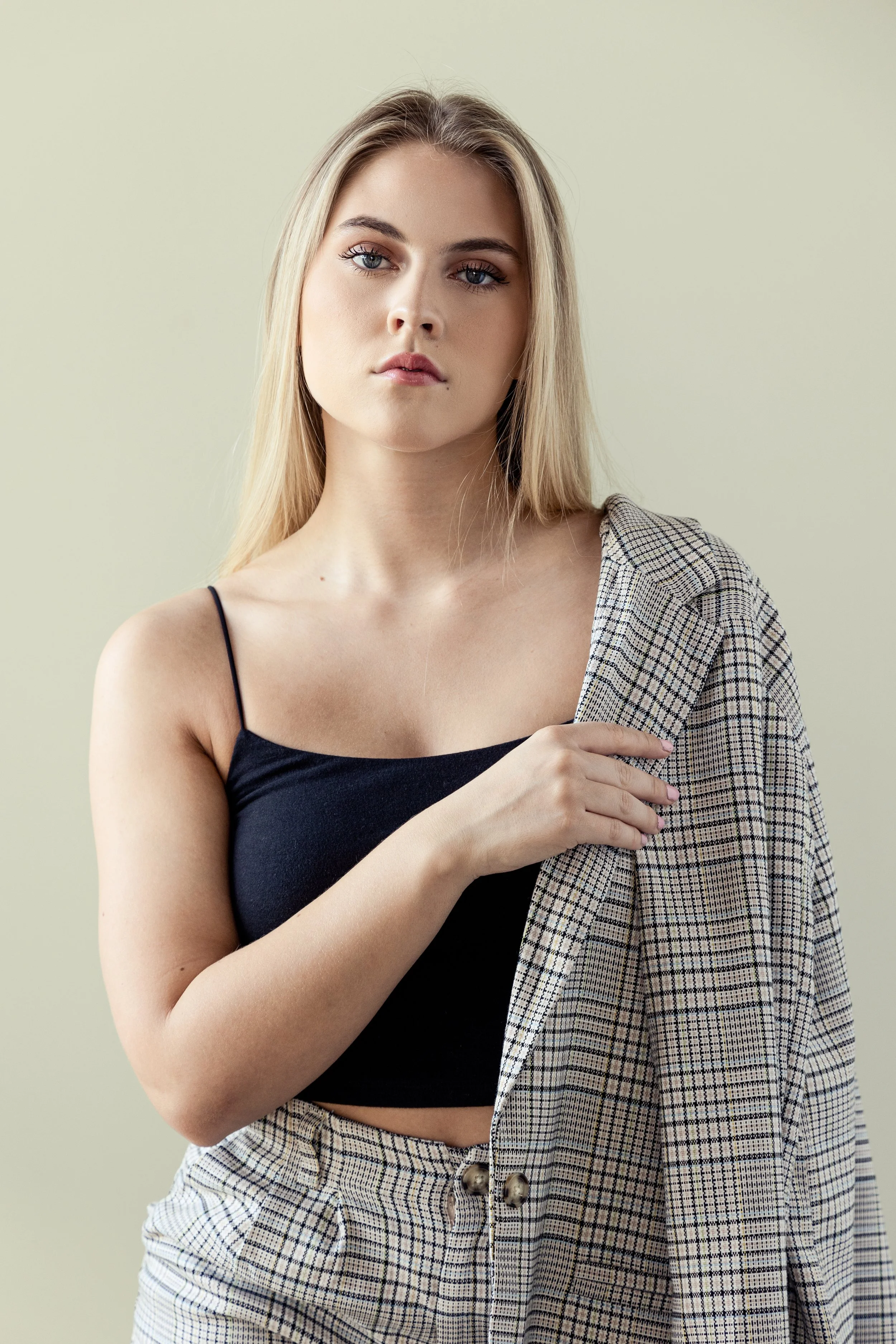 A young woman with blonde hair and blue eyes wearing a black spaghetti strap top and plaid pants, holding a matching plaid blazer over her shoulder, standing against a plain light green background.