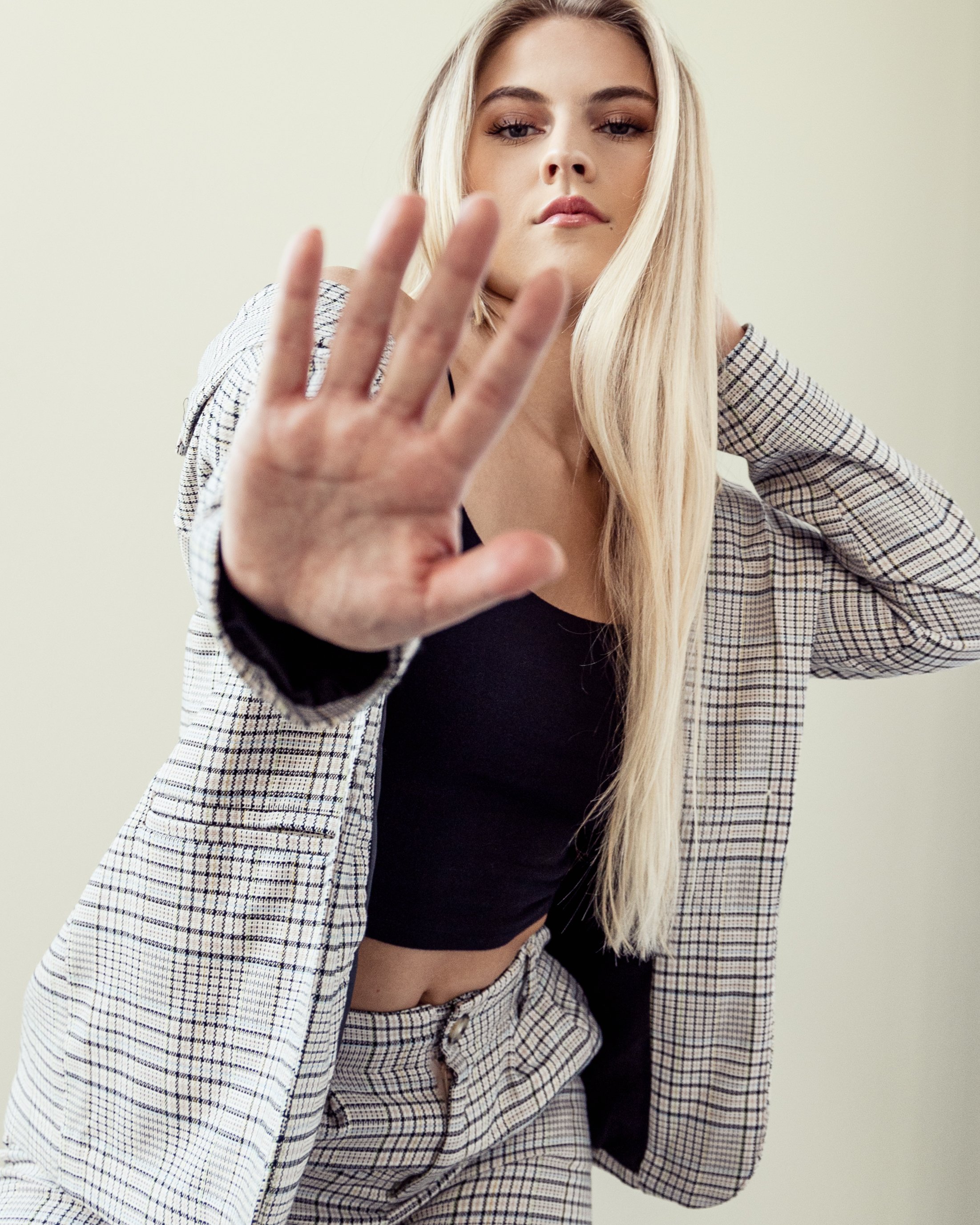 A woman with long blonde hair wearing a plaid blazer and matching pants, black top, holding her hand up with her palm facing the camera as if signaling 'stop'.