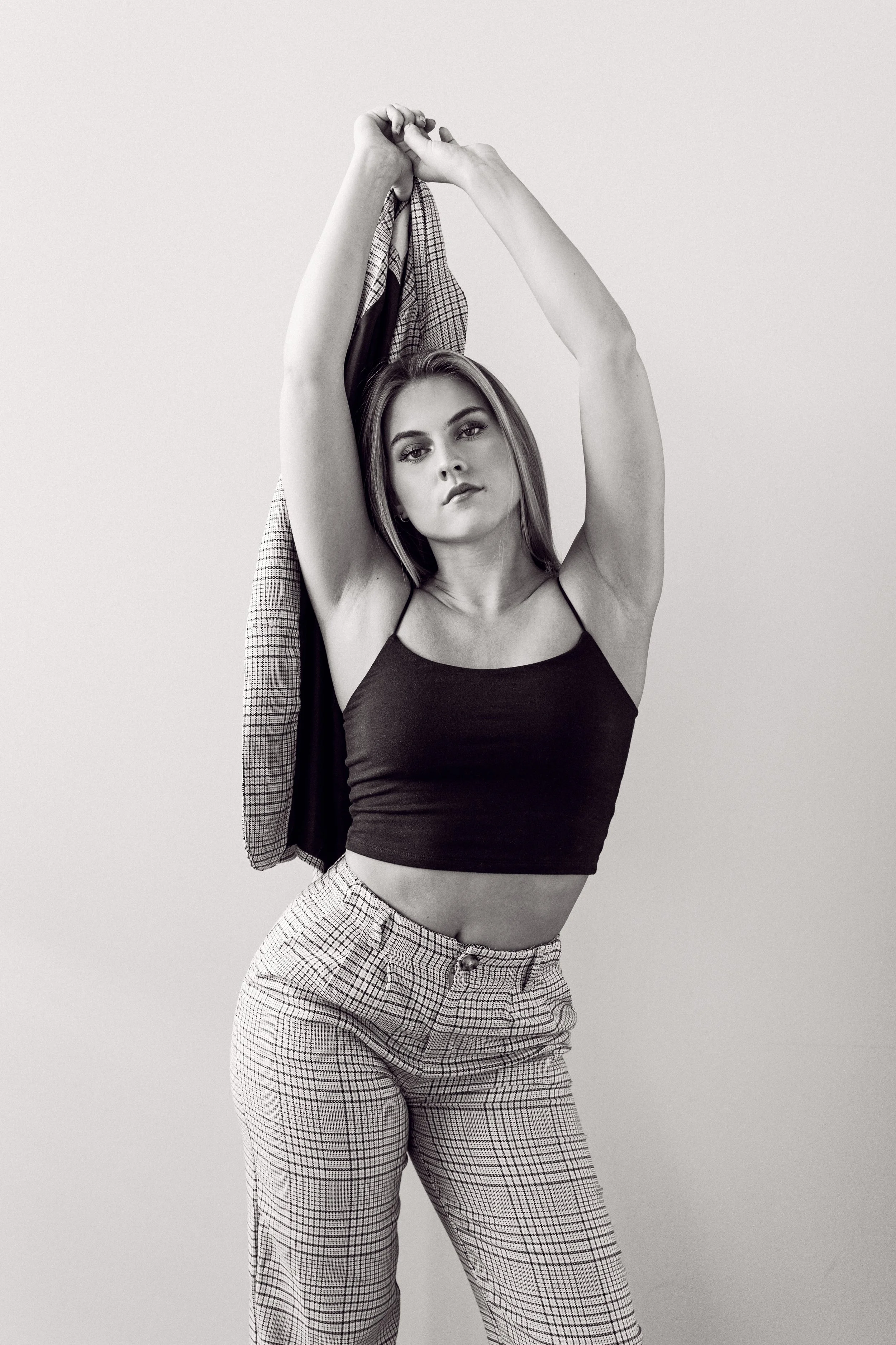 A woman with long hair in a black crop top and plaid pants raising her arms and holding a plaid shirt behind her head, looking at the camera against a plain background.