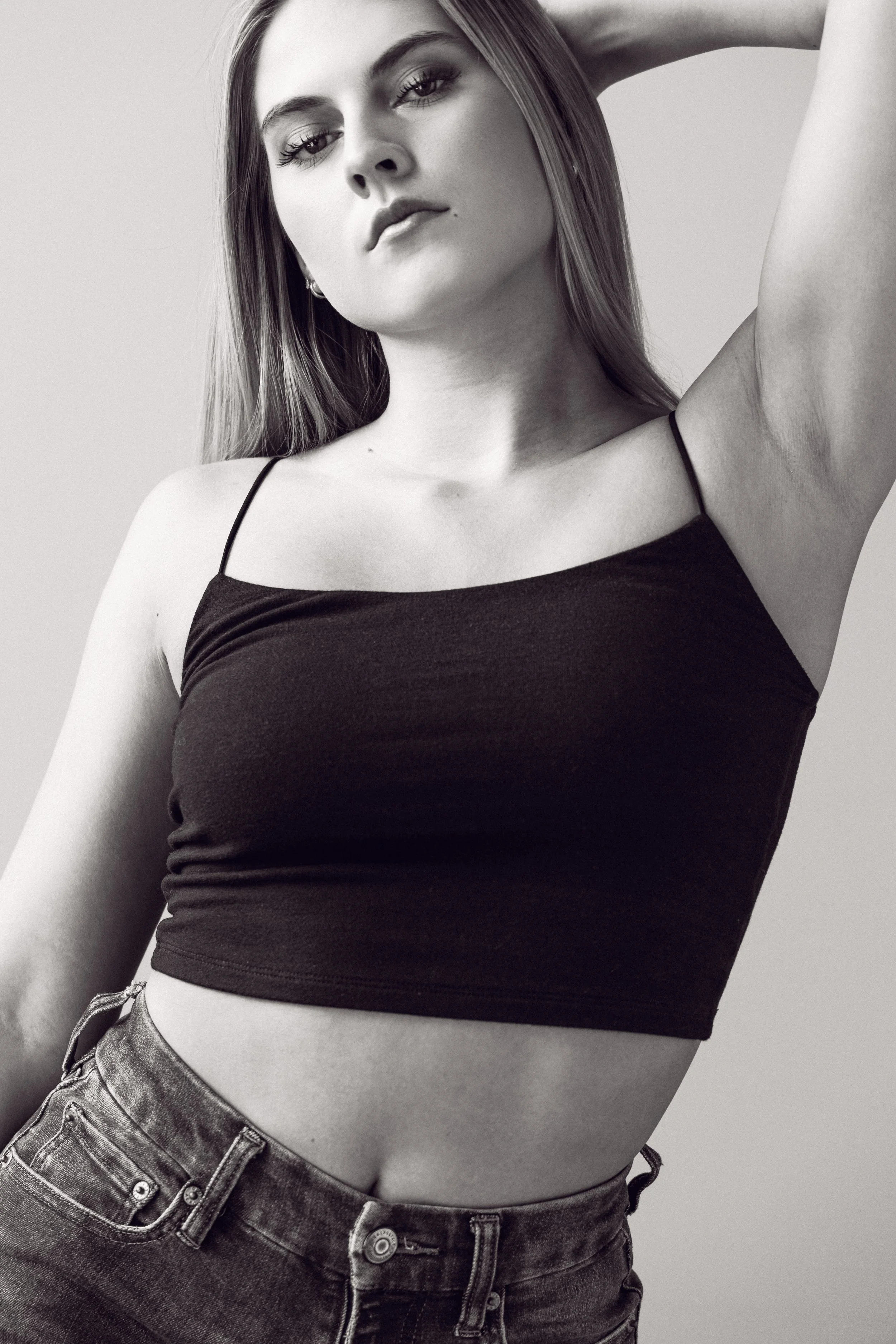 A black and white photograph of a young woman with long, straight hair, wearing a black spaghetti strap crop top and high-waisted jeans, posing with one arm raised behind her head.