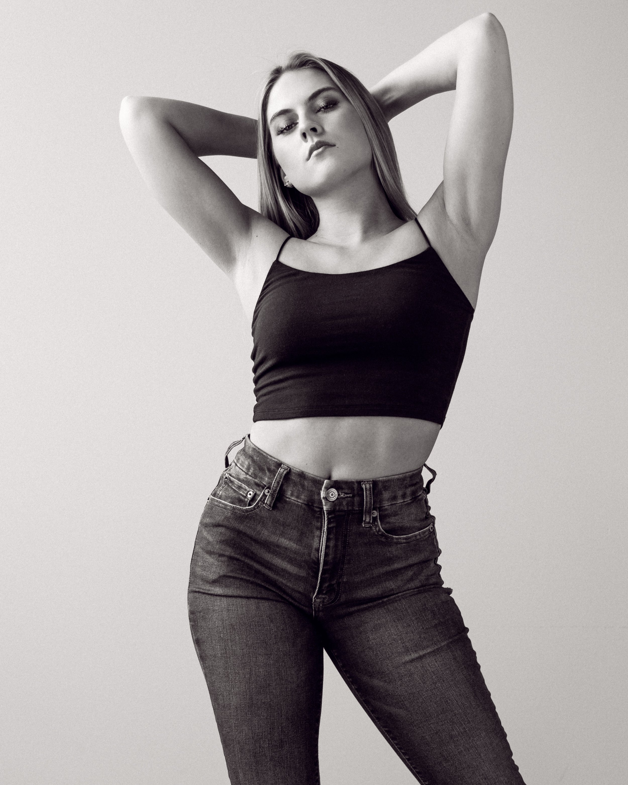 A young woman in a black crop top and jeans posing with her hands behind her head, looking confidently at the camera in a black-and-white photo.