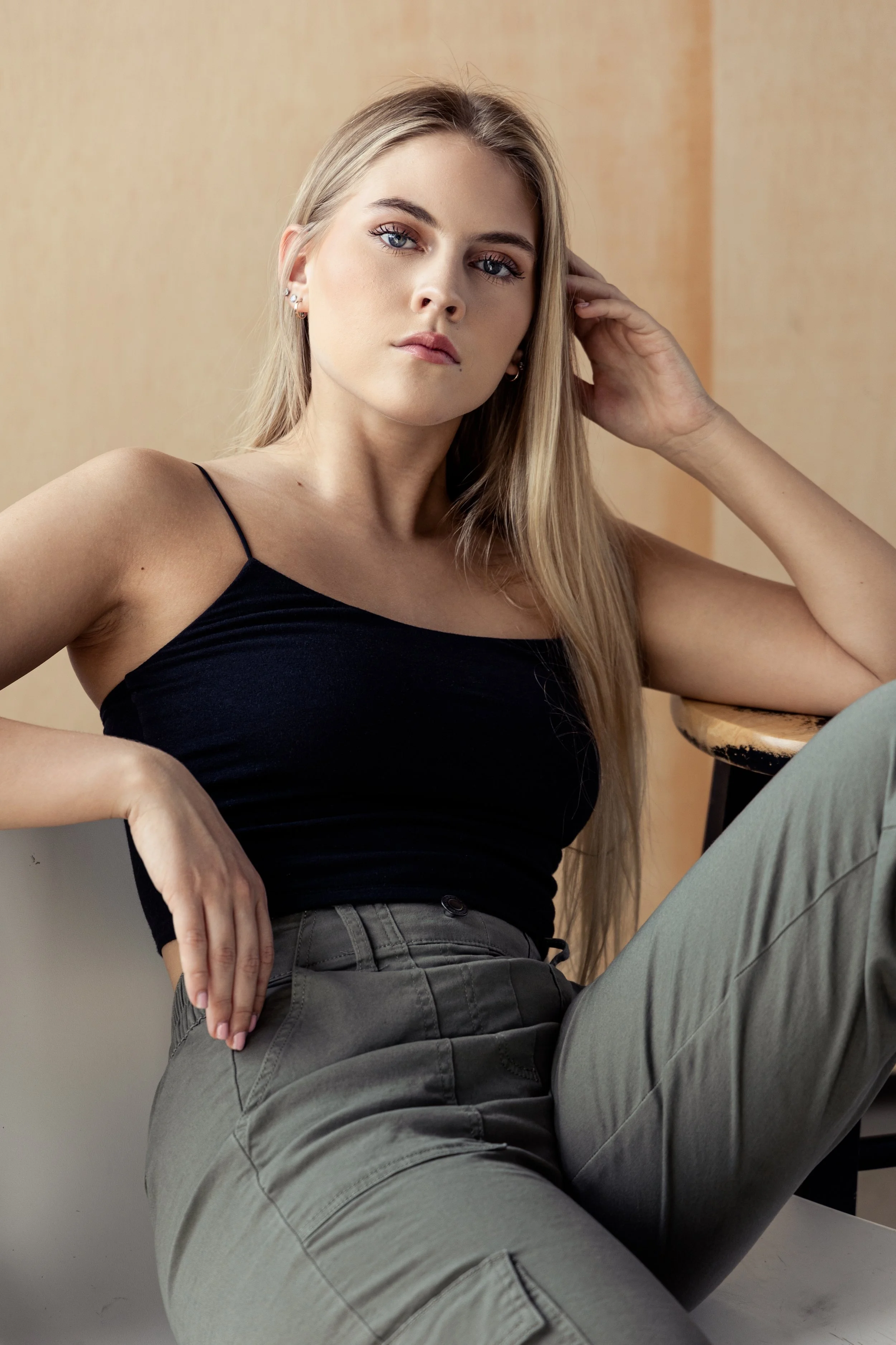 A young woman with long blonde hair wearing a black spaghetti strap top and gray cargo pants, sitting in a relaxed pose with one hand on her head and the other resting on her waist, looking into the camera.