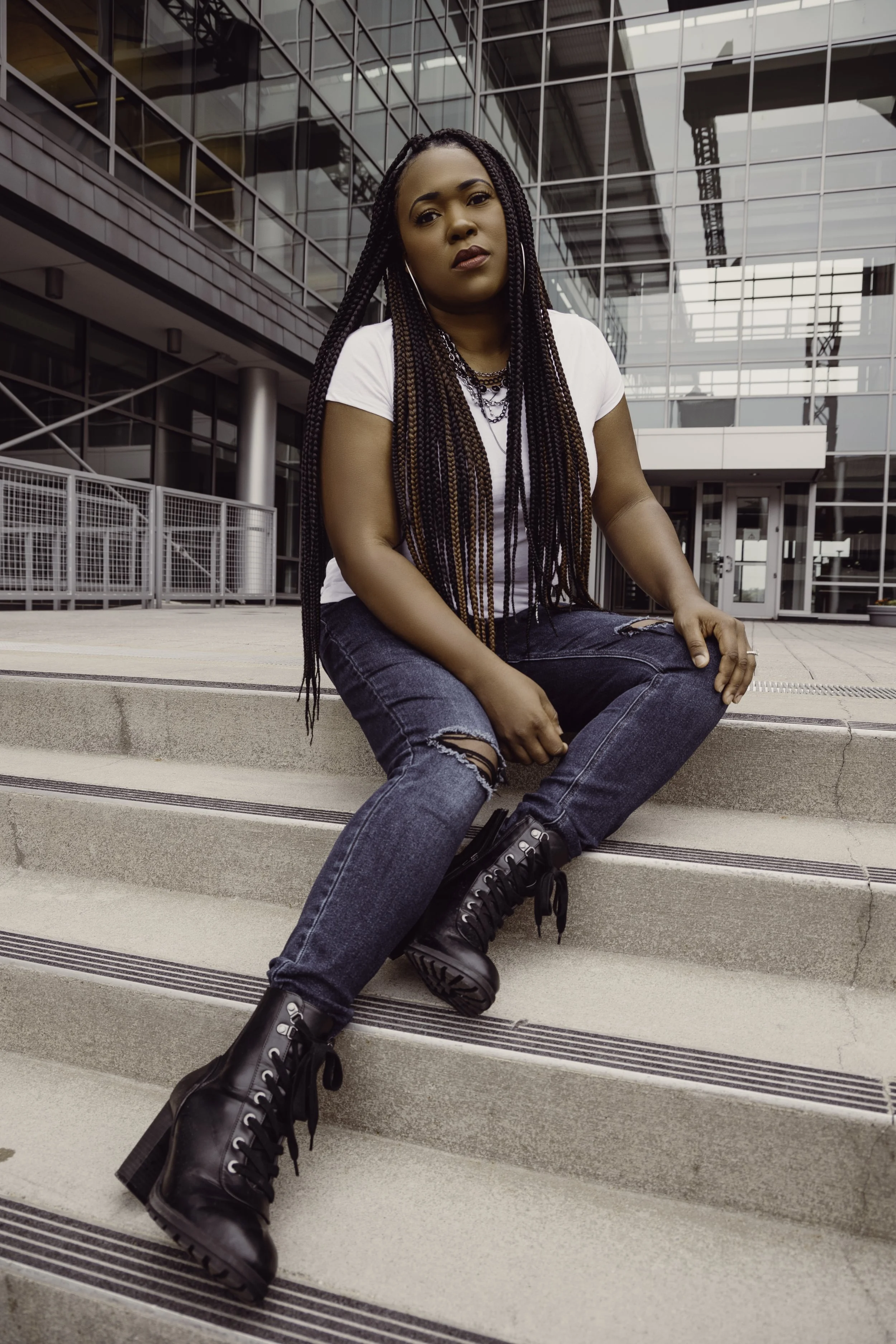 A woman with long braided hair sitting on outdoor stairs in front of a modern glass building, wearing a white t-shirt, ripped jeans, and black lace-up boots.