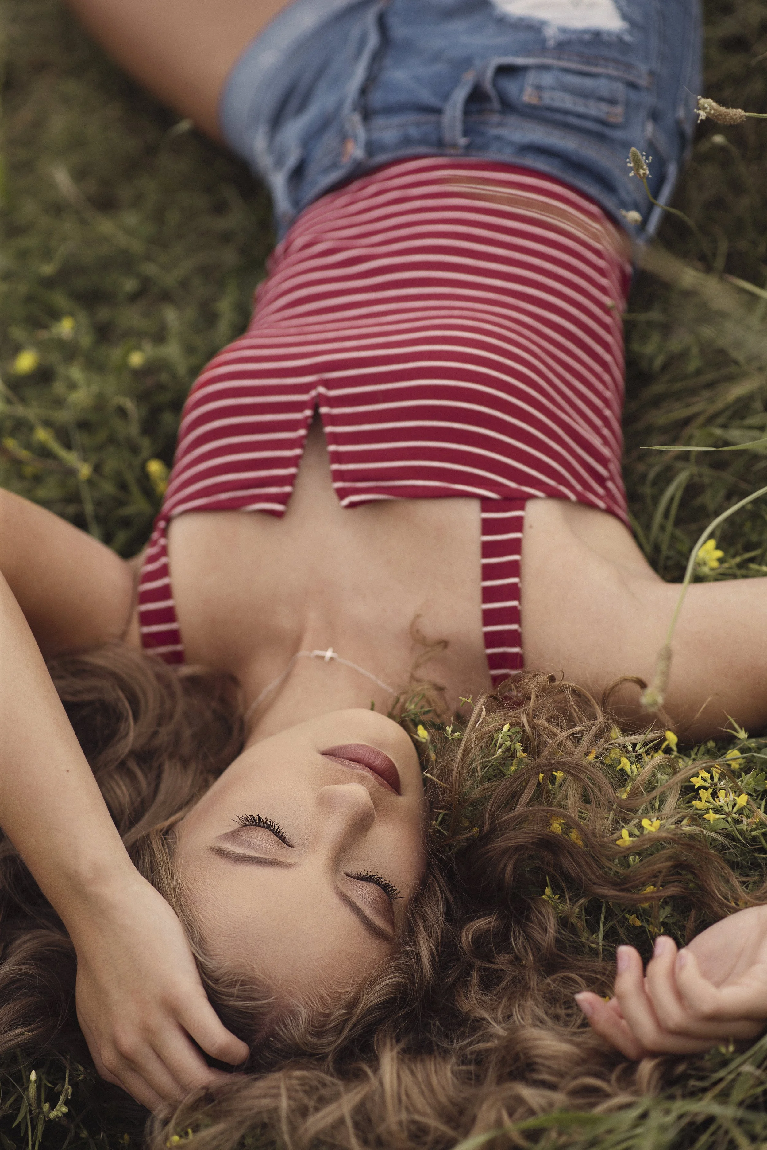 A woman with long curly hair lying on grass with small yellow flowers, eyes closed, wearing a red and white striped sleeveless top and denim shorts.