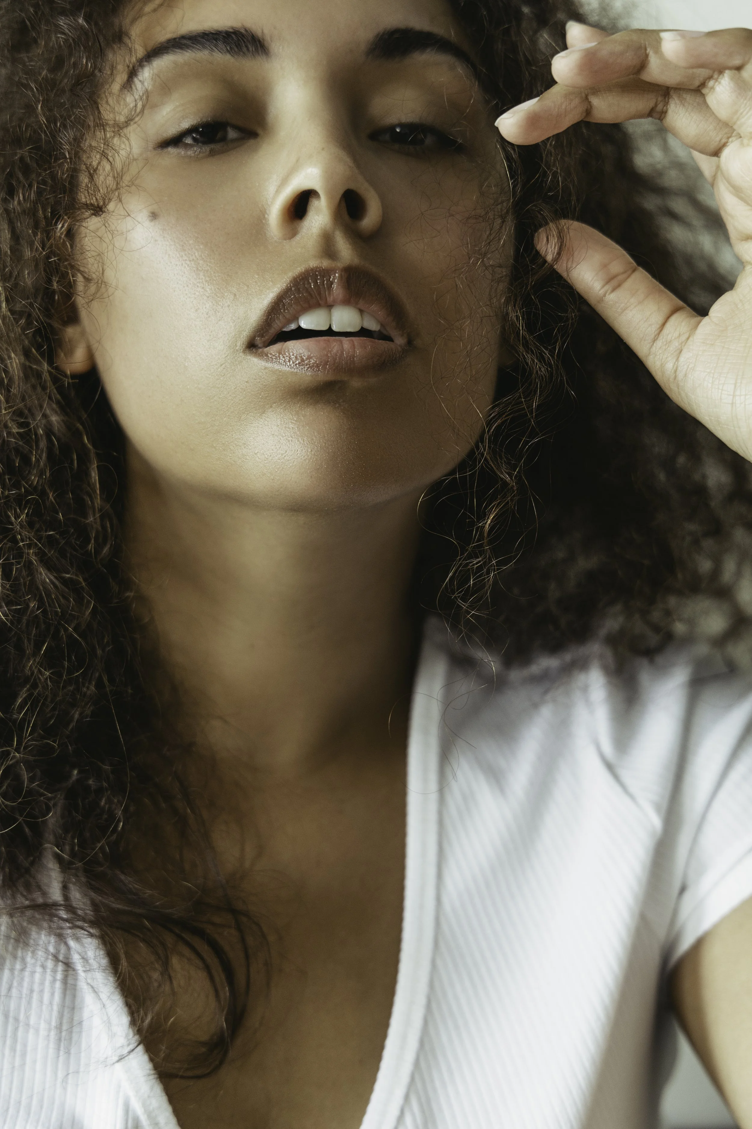 Close-up of a woman with curly hair, partly elevated hand, wearing makeup, slightly open lips, and a white shirt.