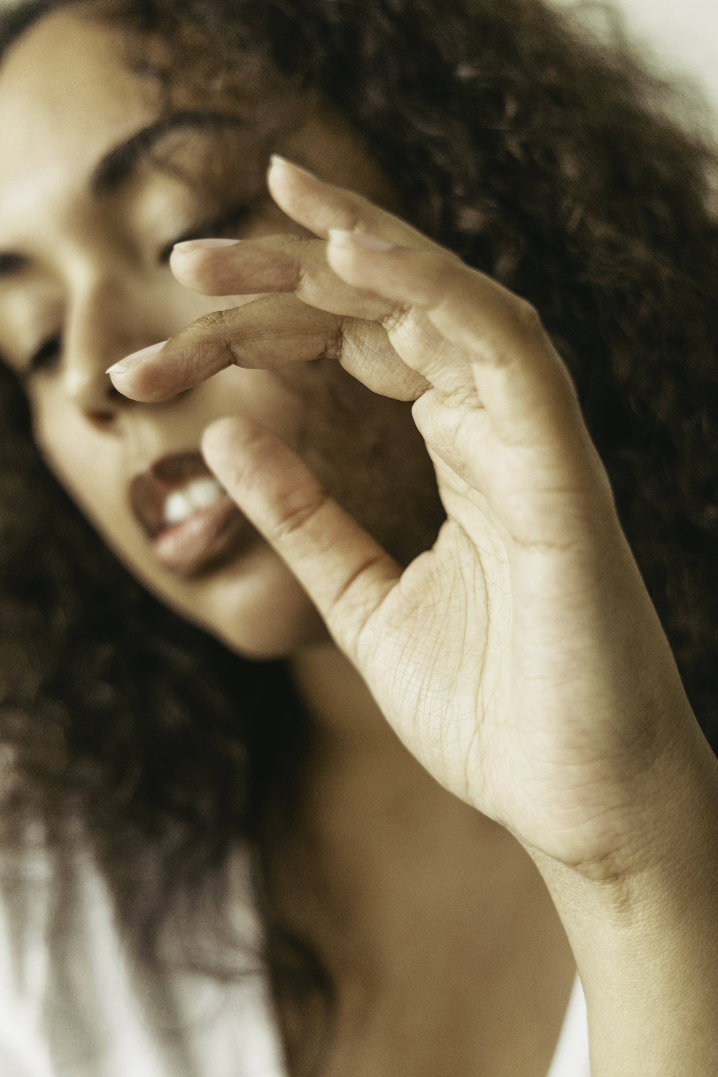 Close-up of a woman with curly hair touching her face with her fingers, eyes closed, and lips slightly parted.