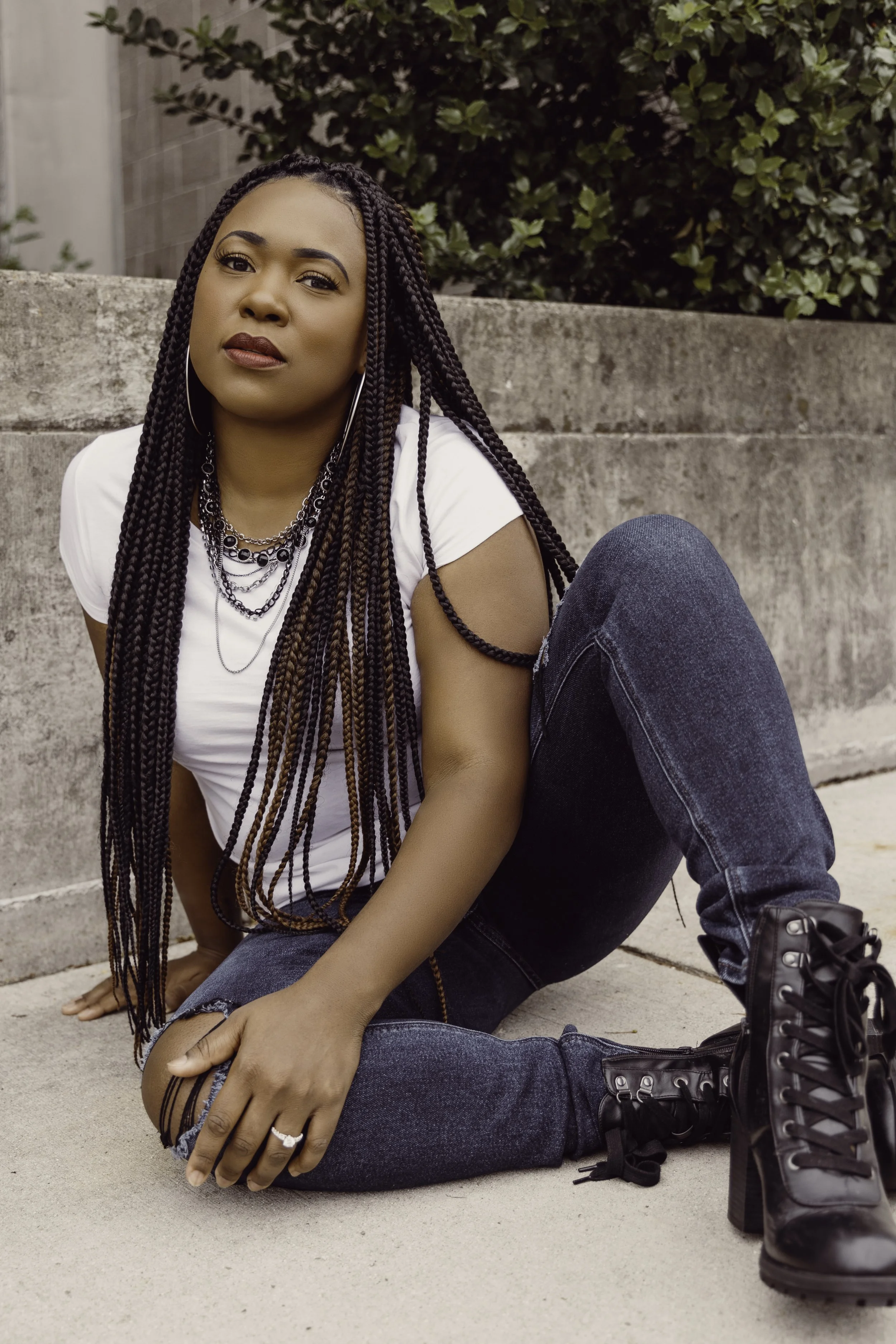 A woman with long braided hair, wearing a white t-shirt, dark jeans, and black boots, sitting on the sidewalk against a concrete wall with greenery in the background.