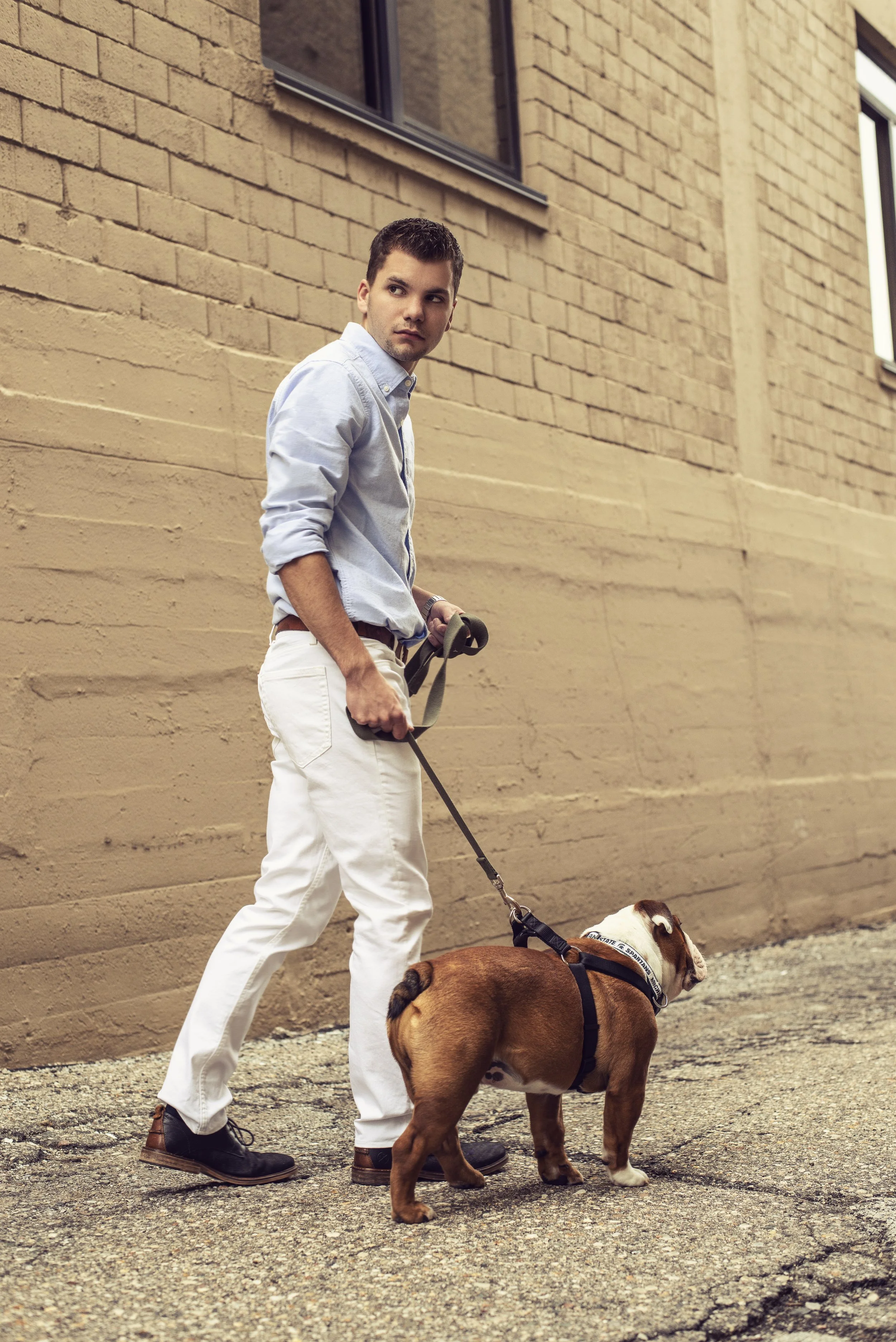 A young man holding a leash with a Bulldog walking beside him along a gravel sidewalk in front of a beige brick wall.
