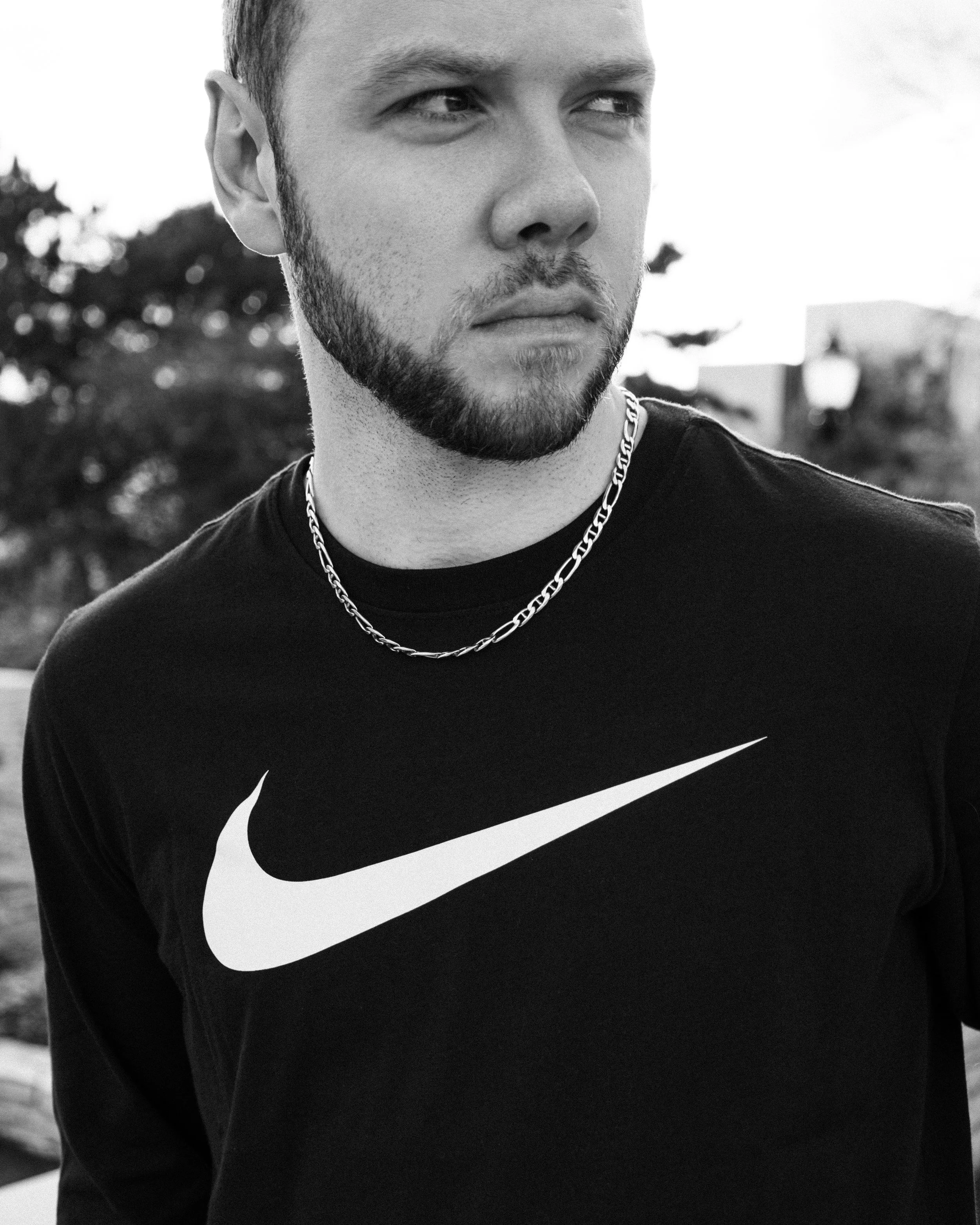 A young man with a beard and short hair looking to the side, wearing a black Nike shirt and a chain necklace, outdoors with trees in the background.