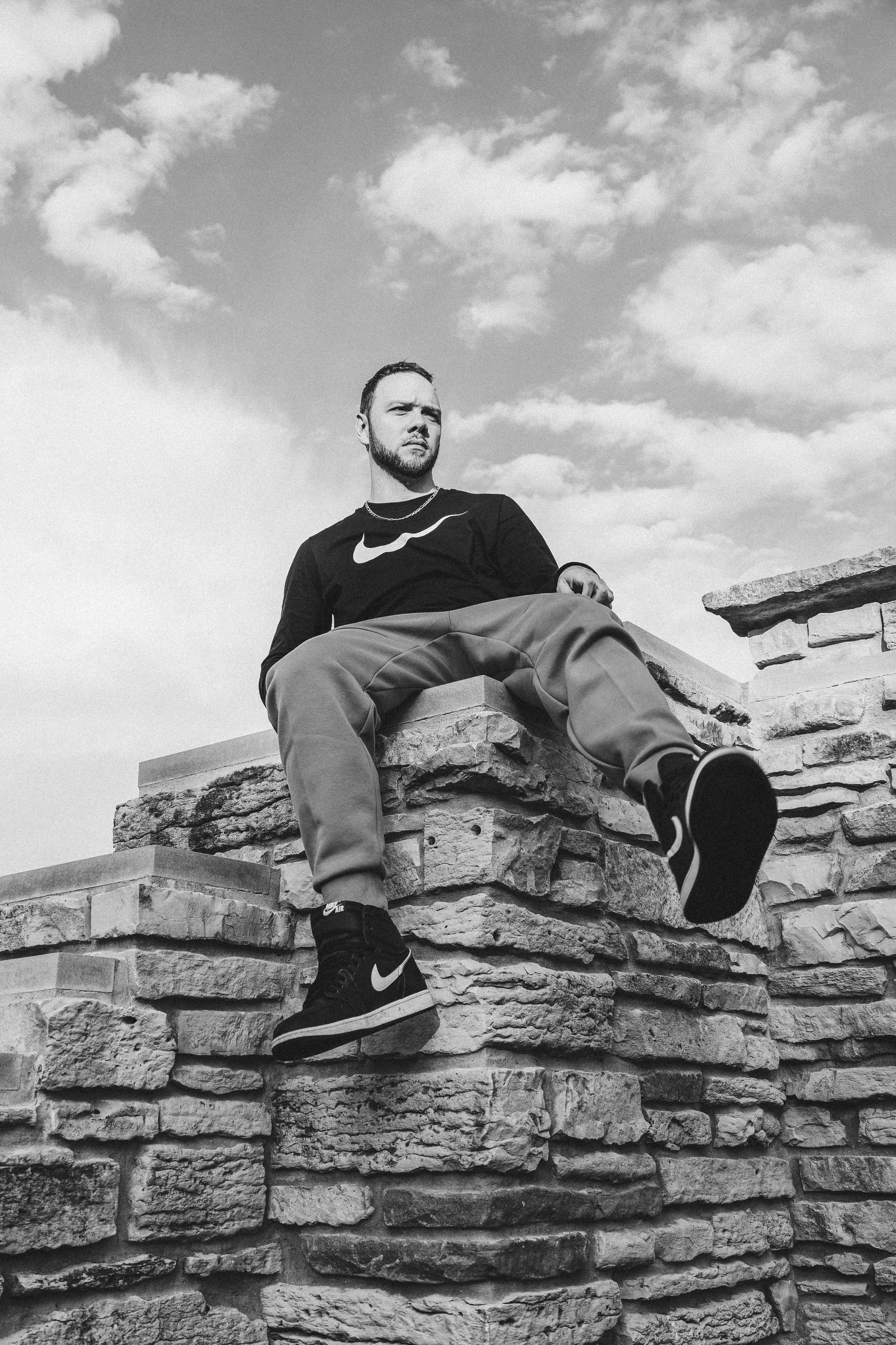 A man sitting on a stone wall under a cloudy sky, wearing a Nike logo t-shirt and Nike sneakers, looking to the side.