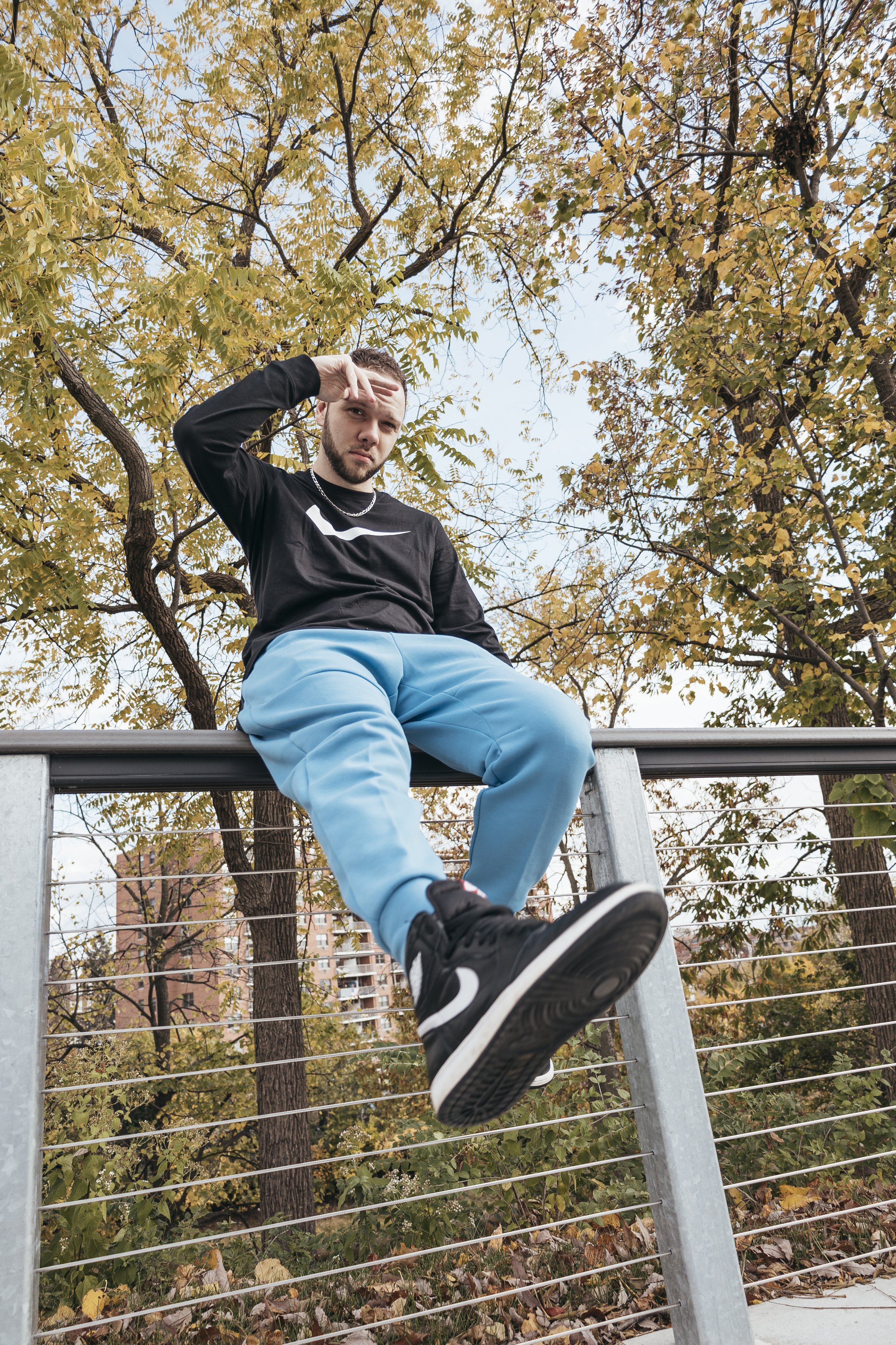 Young man in black Nike shirt and blue pants sitting on a metal railing during autumn, with trees and buildings in the background.