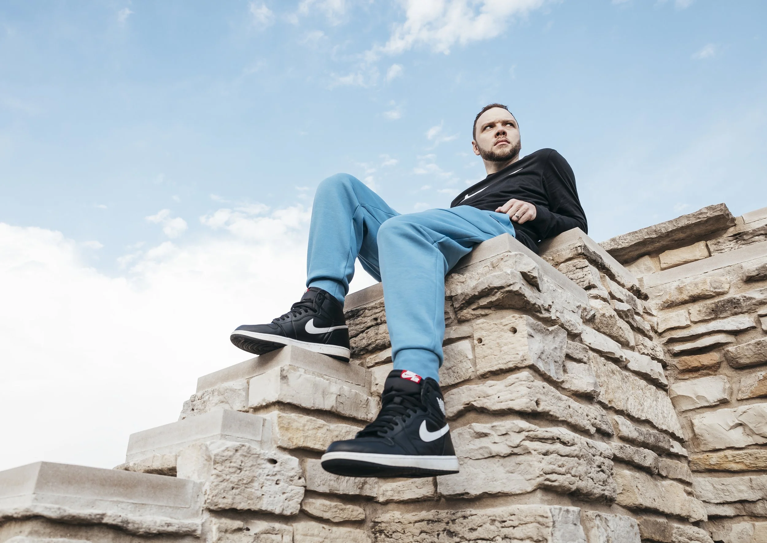A young man with a beard and short hair sitting on a stone wall, wearing black Nike sneakers, black sweatshirt, and blue pants, looking off into the distance against a blue sky with clouds.