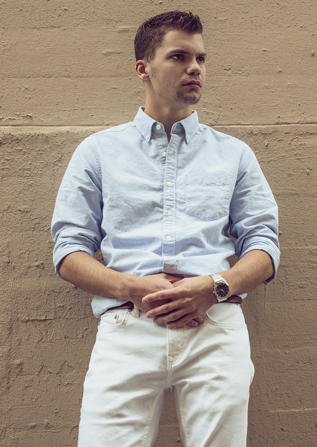 A young man with short hair wearing a light blue button-up shirt with rolled-up sleeves, white pants, and a watch, standing against a beige brick wall.