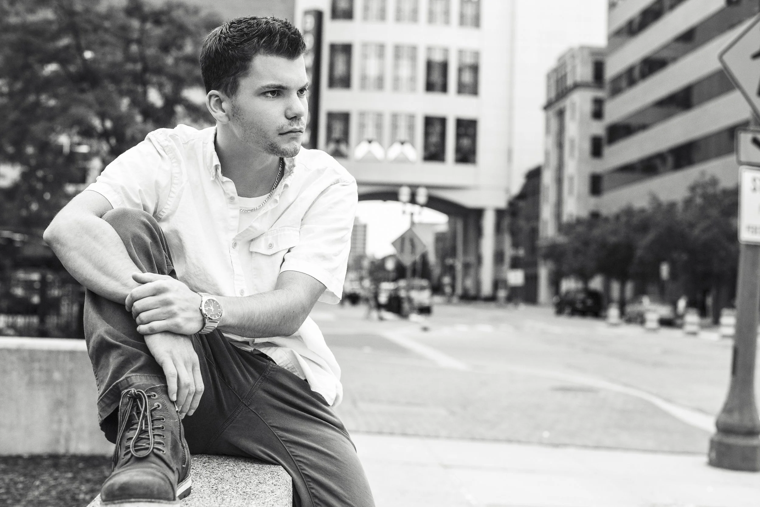 A young man sitting on a low concrete wall in an urban setting, dressed in a white button-down shirt, dark pants, and boots, with a watch and chain necklace, looking thoughtfully to the side.