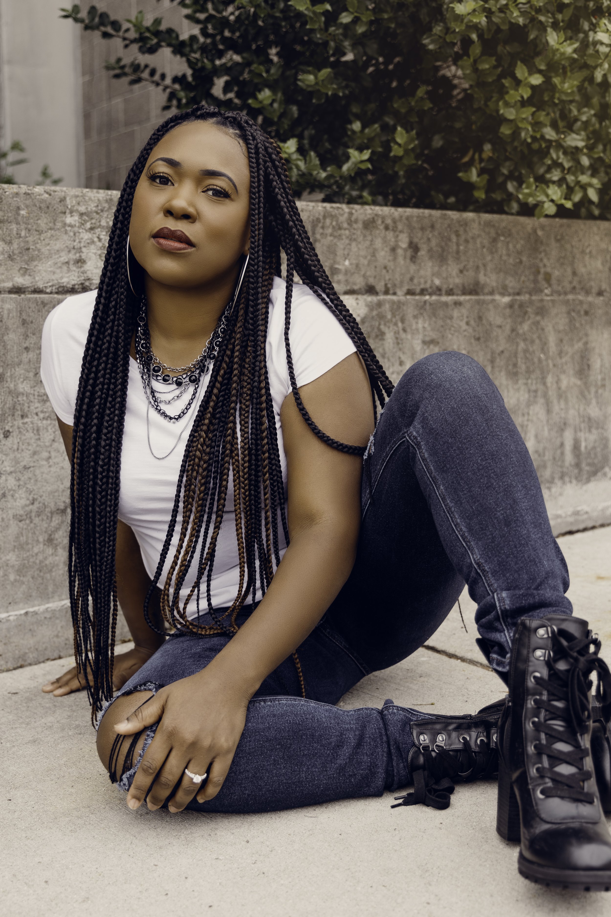 A woman with long braided hair wearing a white t-shirt, dark jeans, and black combat boots, sitting on the sidewalk against a concrete wall outdoors.