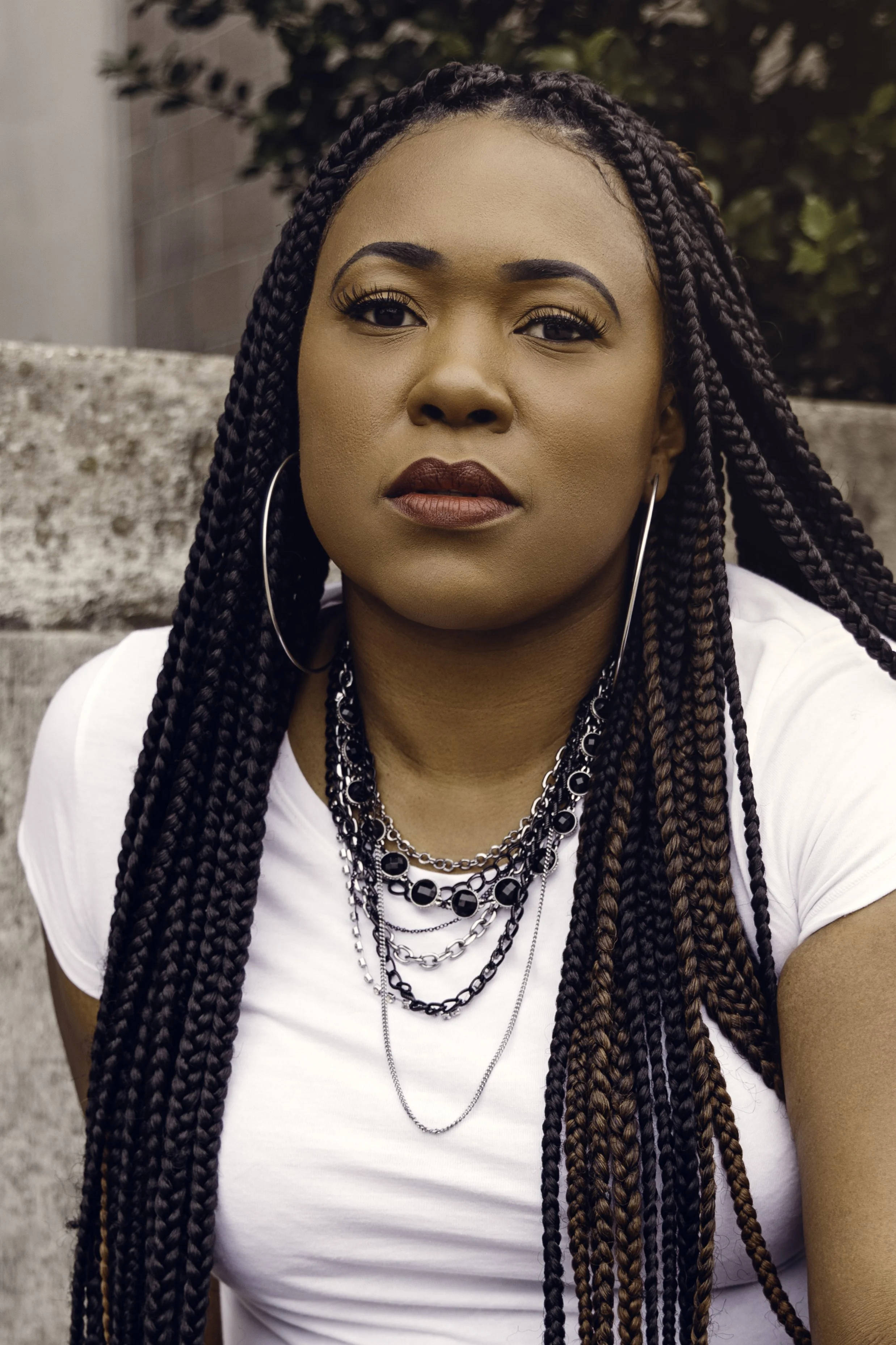 Close-up portrait of a woman with braided hair wearing hoop earrings, multiple layered necklaces, and a white shirt, sitting outdoors against a stone and foliage backdrop.