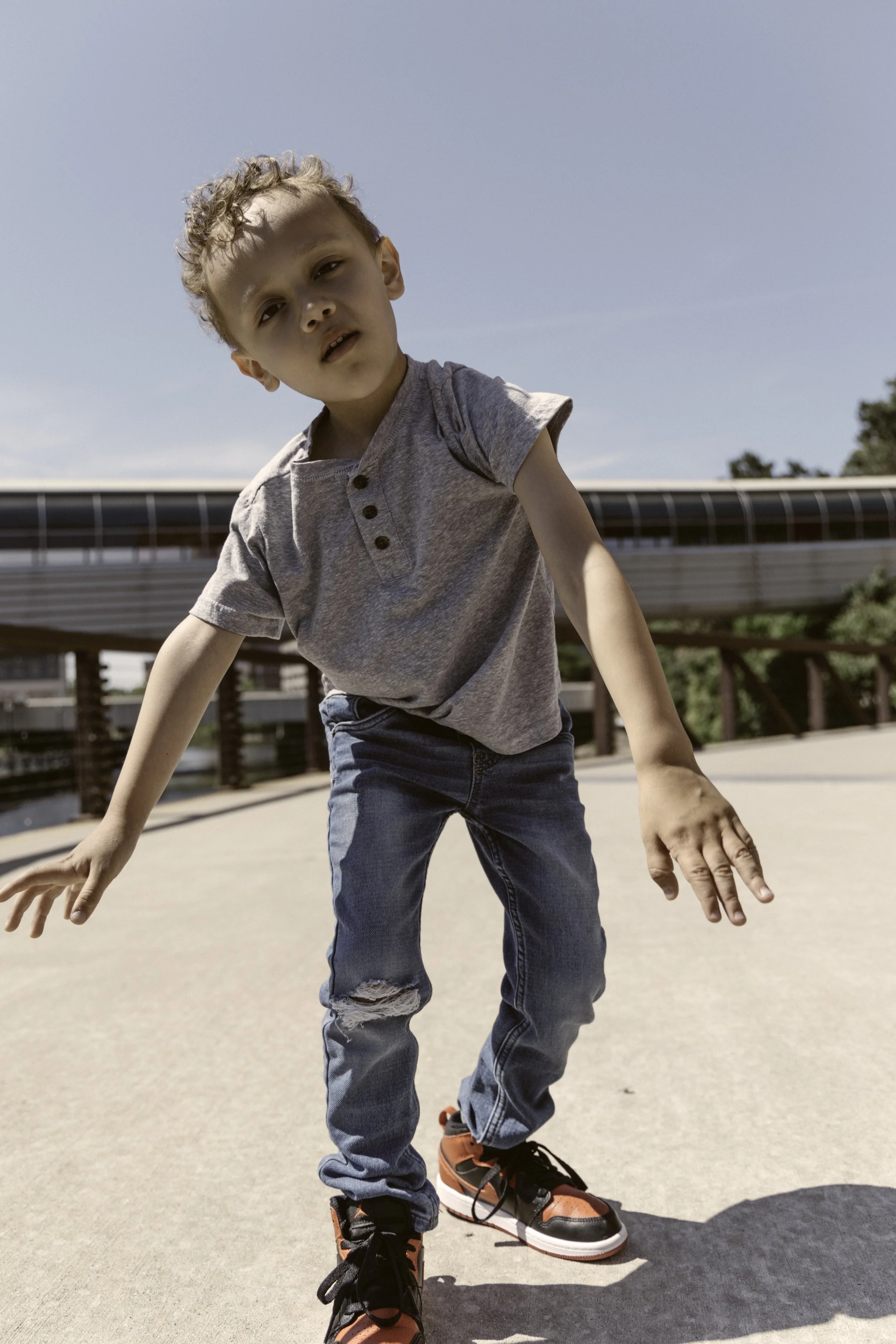 Young boy with curly hair wearing a gray t-shirt, ripped jeans, and red and black sneakers, leaning forward on an outdoor walkway on a sunny day.