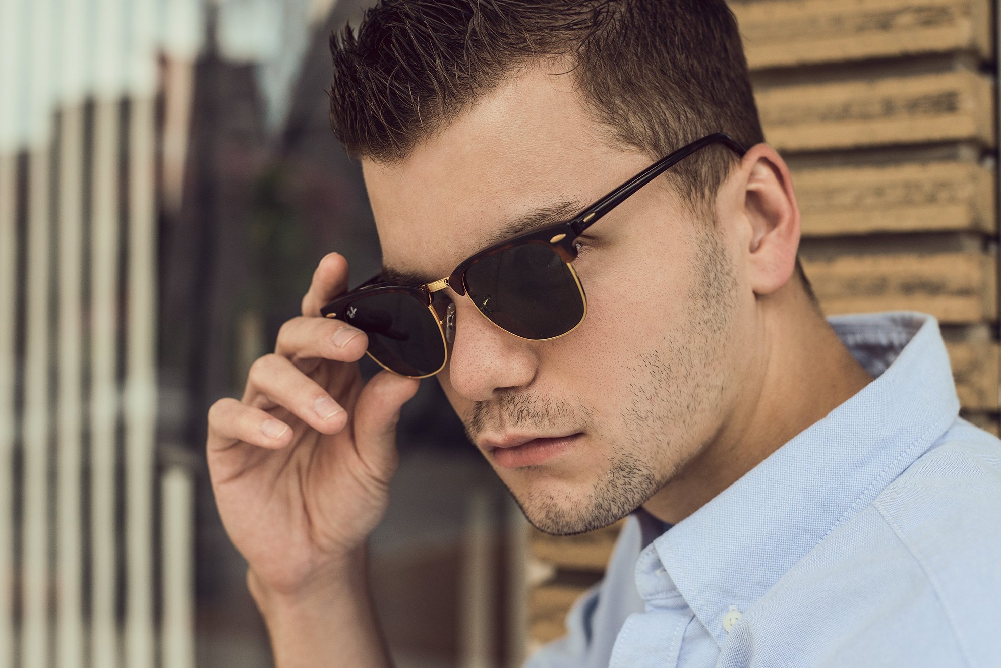 A young man with short brown hair and stubble, wearing sunglasses and a light blue button-up shirt, is adjusting his sunglasses with his right hand.