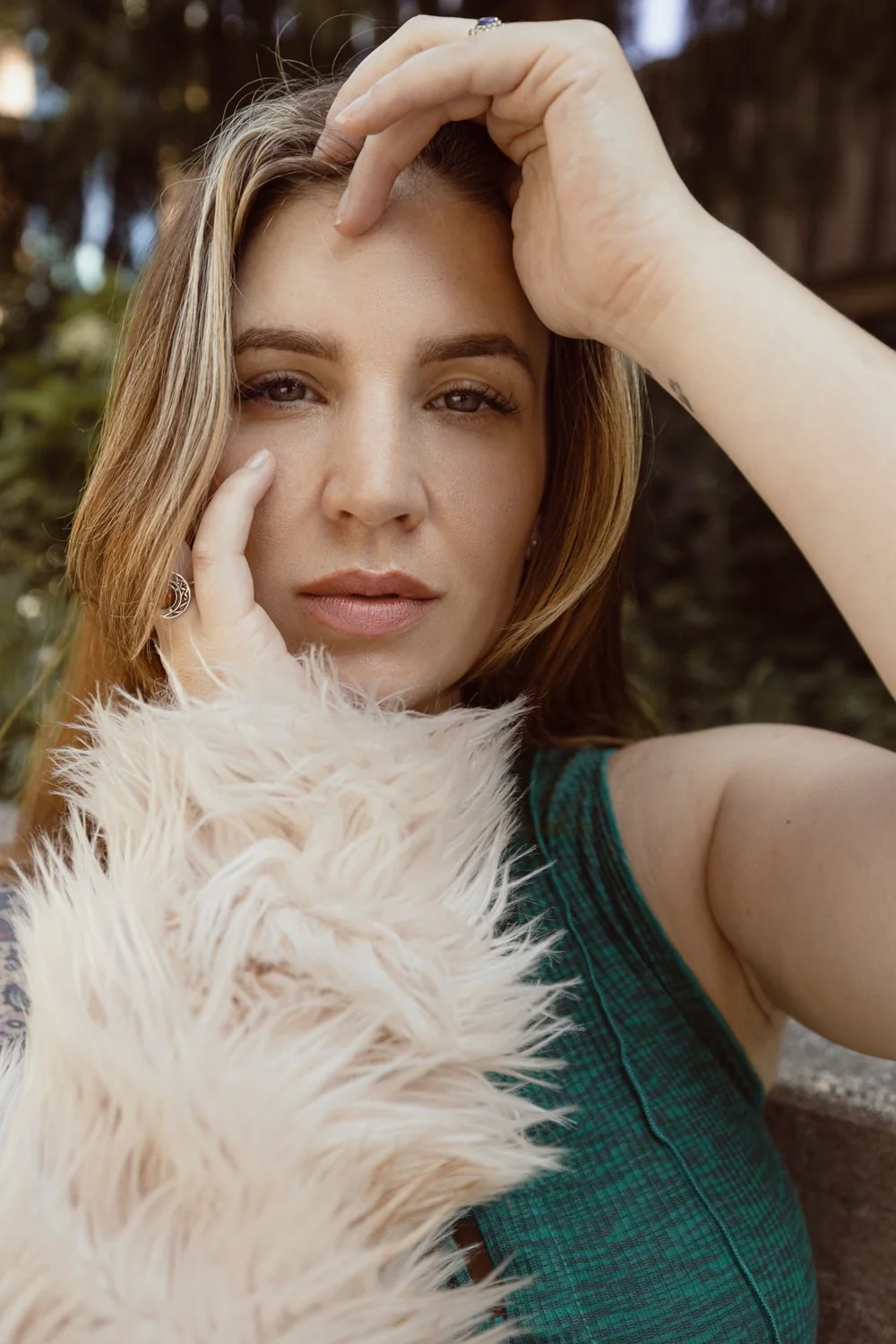 Close-up of a woman with light brown hair, resting her hand on her forehead, wearing a teal checkered top and a fluffy cream-colored scarf, outdoors with blurred greenery in the background.