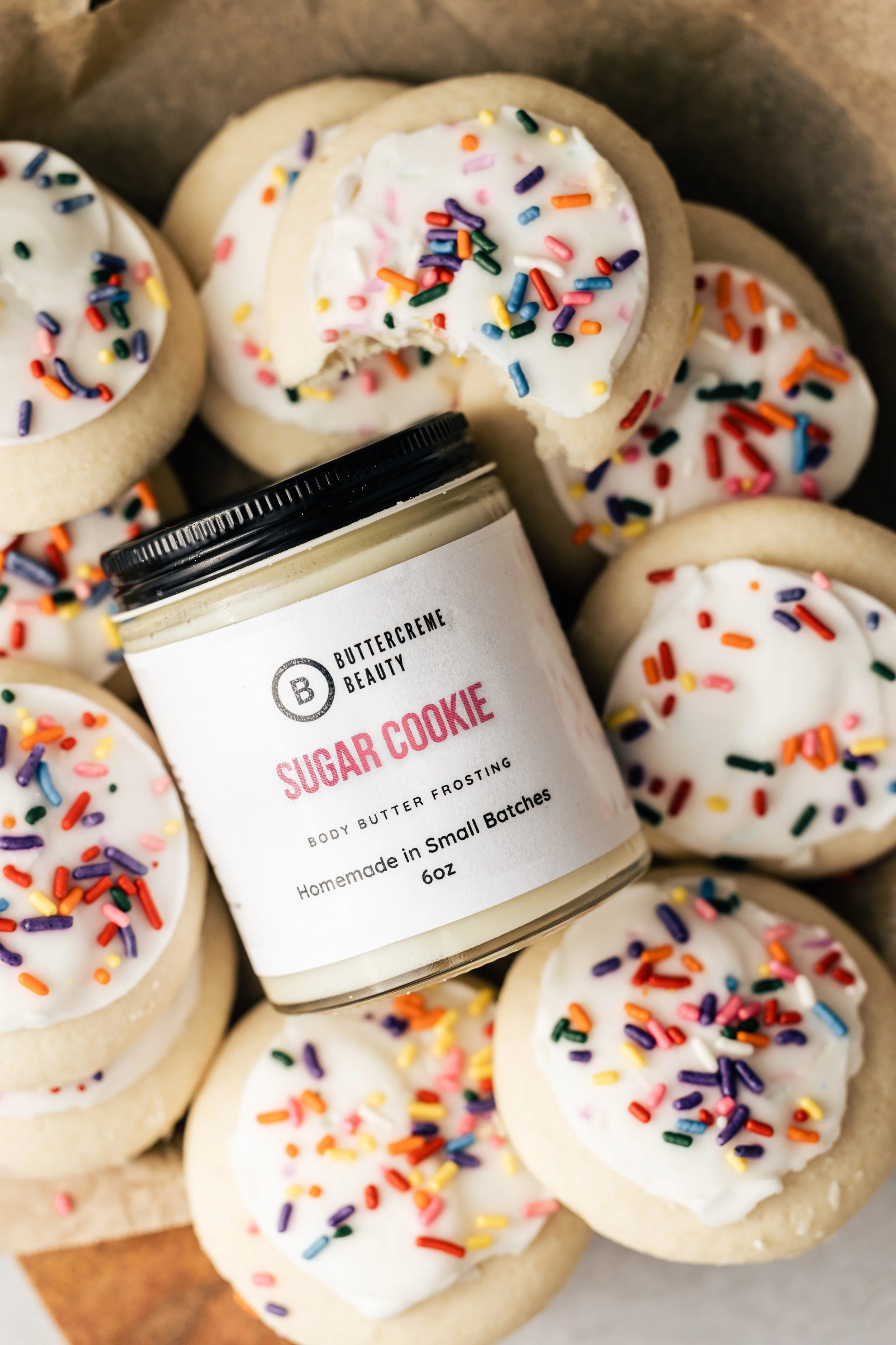 Assorted sugar cookies with white icing and colorful sprinkles surrounding a jar labeled 'Sugar Cookie' body butter frosting.
