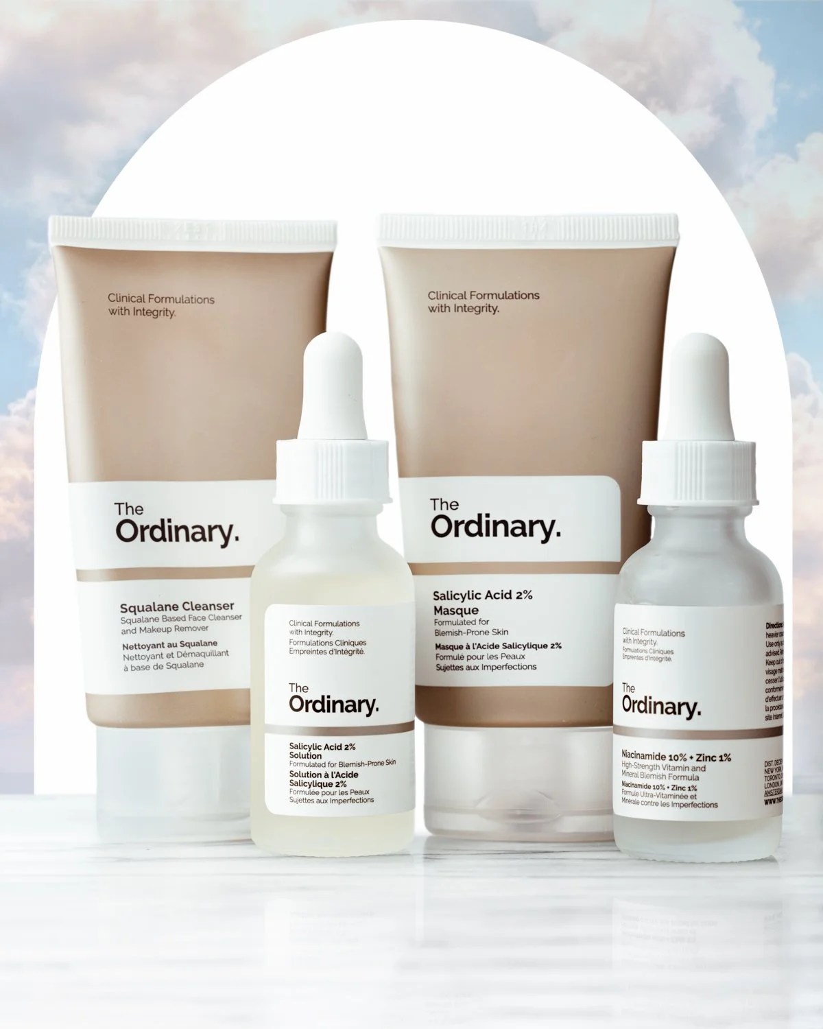Skincare products from The Ordinary, including Squalane Cleanser, Salicylic Acid 2% Masque, and Salicylic Acid 2% Solution, arranged on a white surface with a soft sky background.