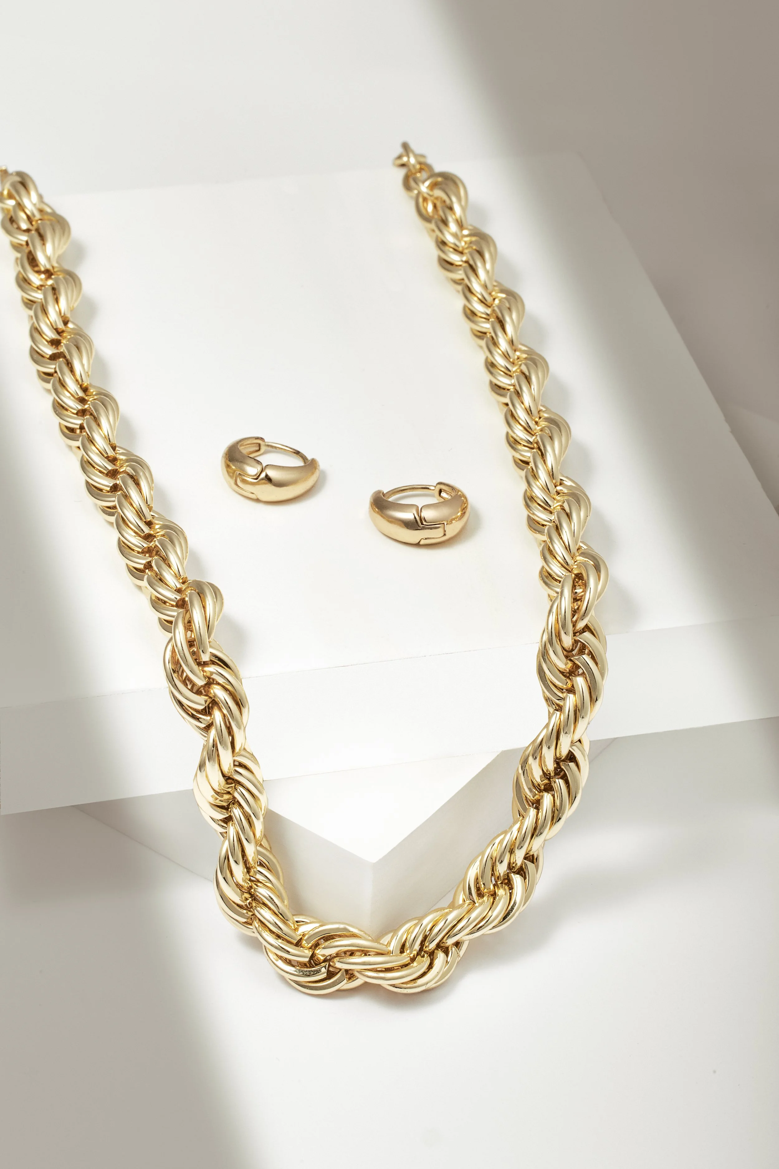 Gold jewelry set comprising a chunky twisted gold chain necklace and matching gold hoop earrings on a white display stand.