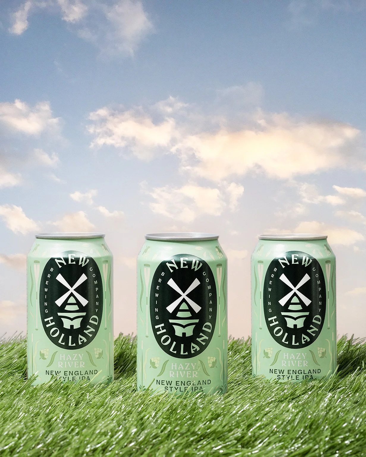 Three cans of Hazy River New England Style IPA by Cold Harbor Brewing Company placed on green grass with a partly cloudy sky background.
