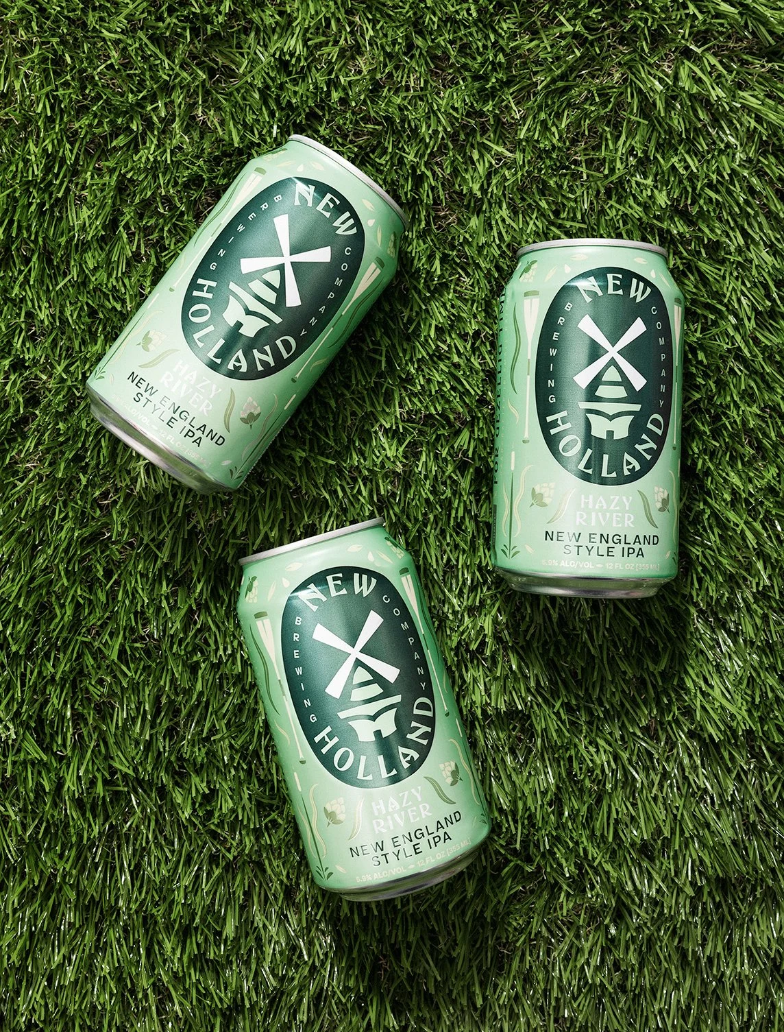 Three cans of Holland Hazy River New England Style IPA beer lying on green grass.